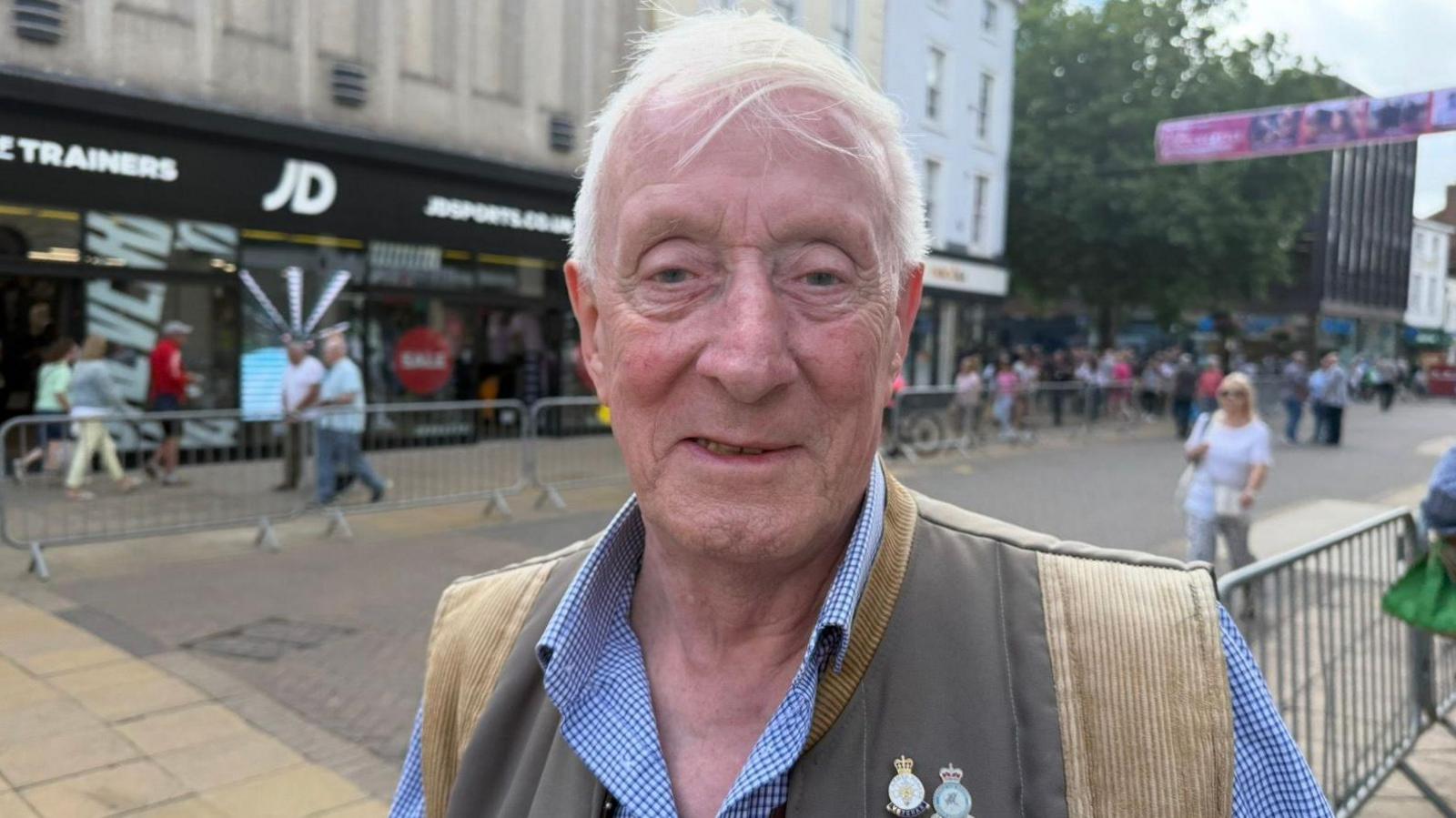 The photo shows a man smiling at the camera. He has white hair, and is wearing a gilet and a blue and white checked shirt. He's also wearing two pin badges with the RAF crest on them. He's standing on a high street, and there are various shops and people behind him. 
