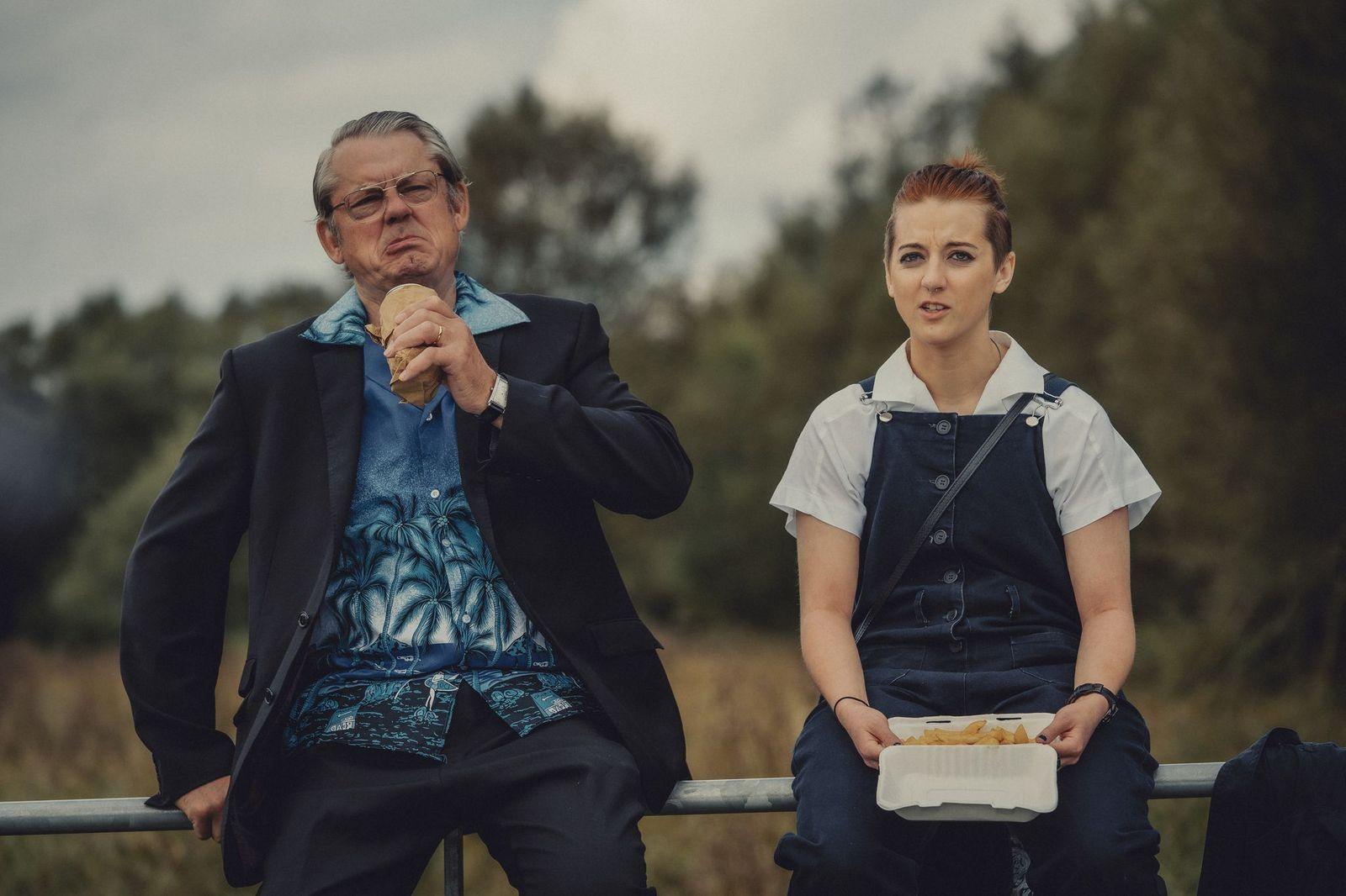 Two characters in the film sitting side by side. On the left is an older man who is looking thoughtful while eating. He is wearing a black suit with a Hawaiian-style blue shirt underneath. The younger woman sitting to his right has red hair pulled back into a ponytail. She is wearing a white, short sleeved shirt and blue dungarees. She is holding a box of chips.