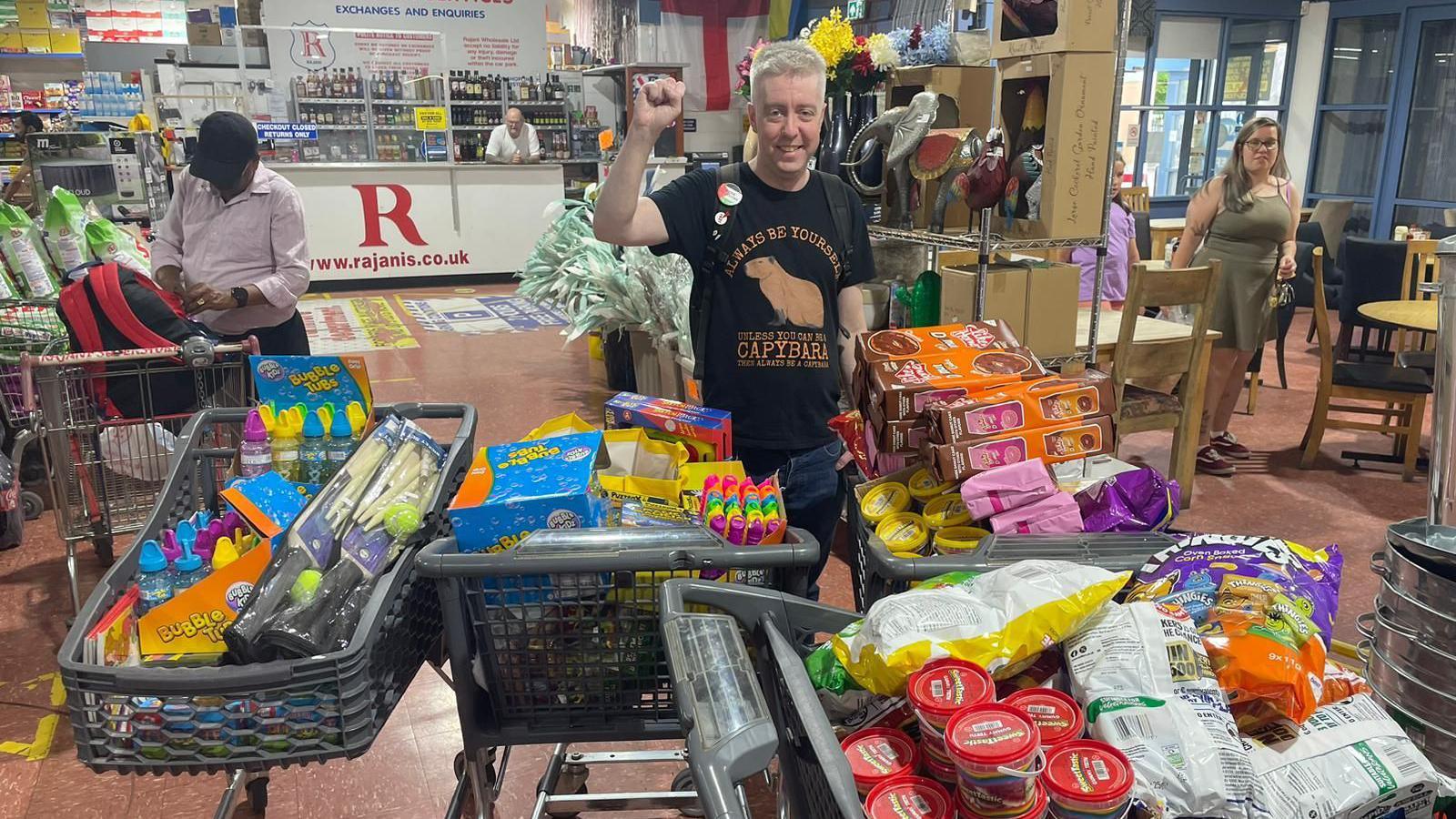 A volunteers stands in a supermarket with his fist raised, in front of trolleys full with sweets and toys. 