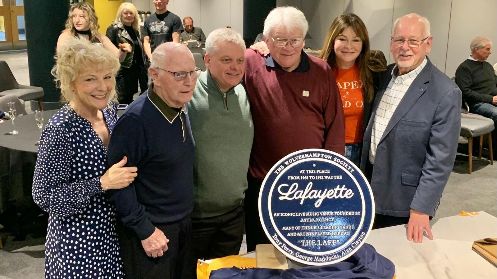 Six people gathered around a blue plaque. They are at an event and there are people standing around drinking prosecco. The plaque say Lafayette and has the branding of the Wolverhampton Society. 