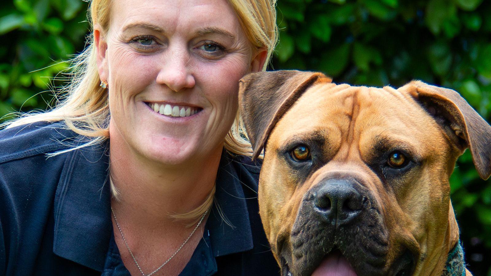 Sam Garvey is smiling at the camera. She is crouched next to a large, brown dog. She has long blond hair and is wearing a blue top