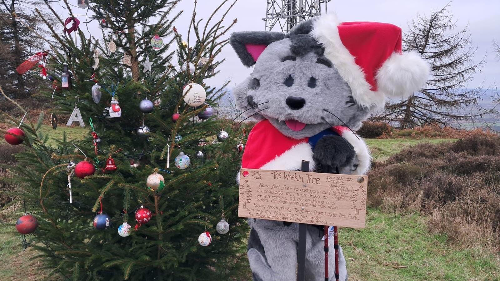 A large grey cat mascot in a santa costume, standing behind a brown sign and a green Christmas tree with baubles hanging from it