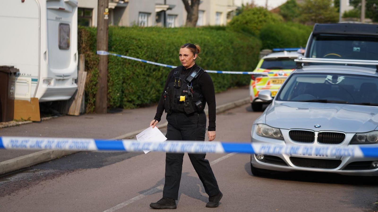A police officer is walking behind the cordon. There is a police car and a silver car behind her and a caravan can be seen parked in the driveway of one of the houses on the road. She is holding a piece of paper.