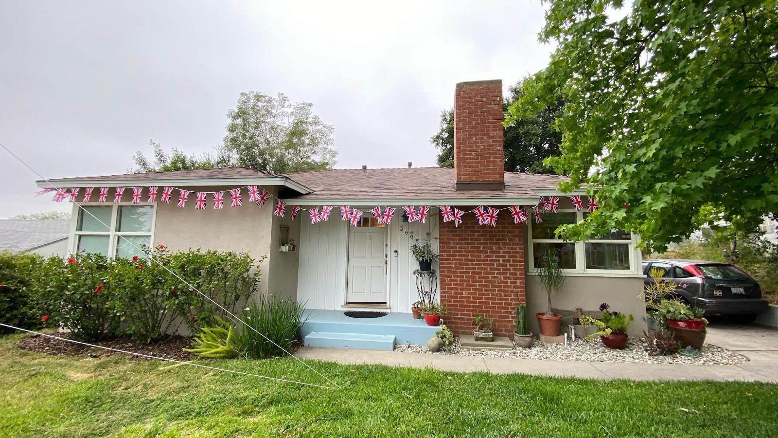 The single-storey home seen in the summer with Union flag bunting decorating the edge of the roof. There is a lawn and plants in pots and there is a car parked to one side.