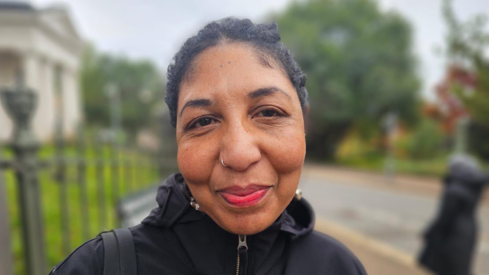 Porsche is smiling at the camera, she had red lipstick on and a silver nose ring. She has dark black hair and is wearing a black coat which is zipped up to her chin.