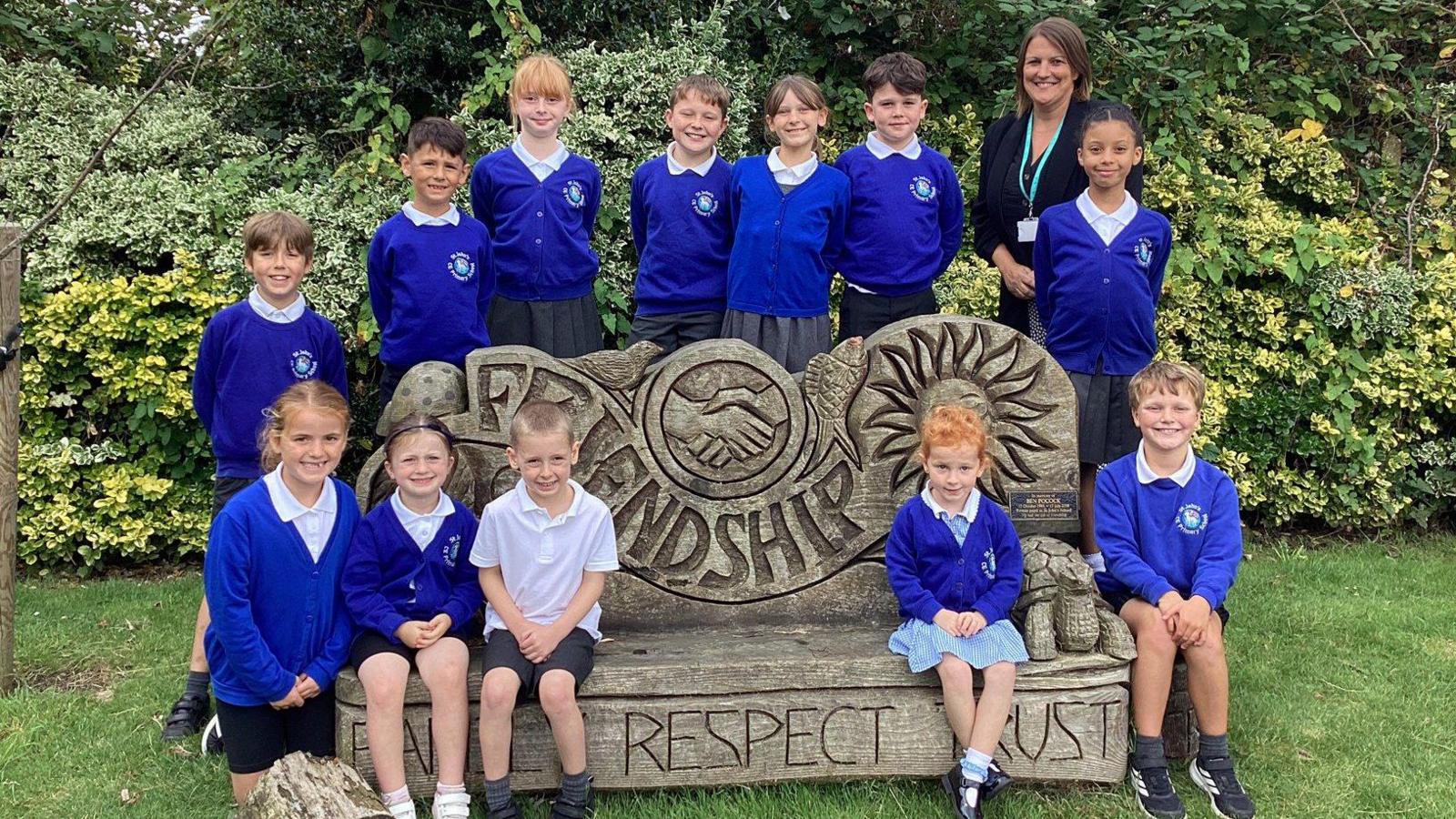 A group of children at St John's Primary in Keynsham sit around a wooden ornament with slogans on it. They are wearing dark blue uniform jumpers and blazers and their teacher is standing with them wearing a black jacket