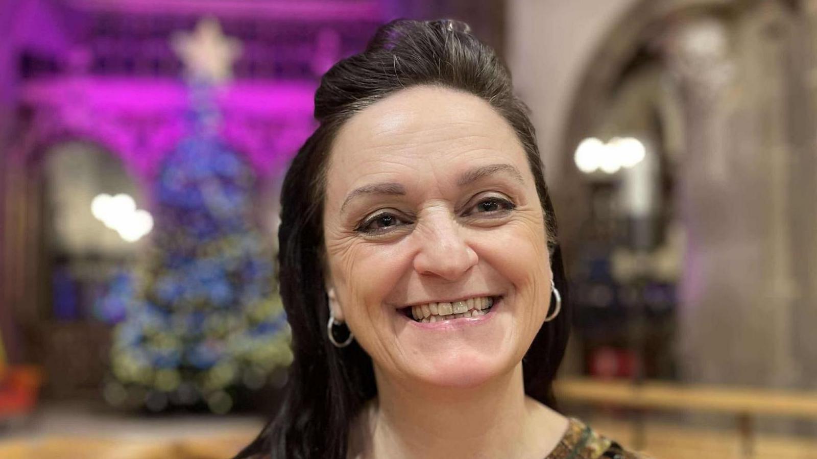 A smiling woman inside Leicester Cathedral with a Christmas tree behind her