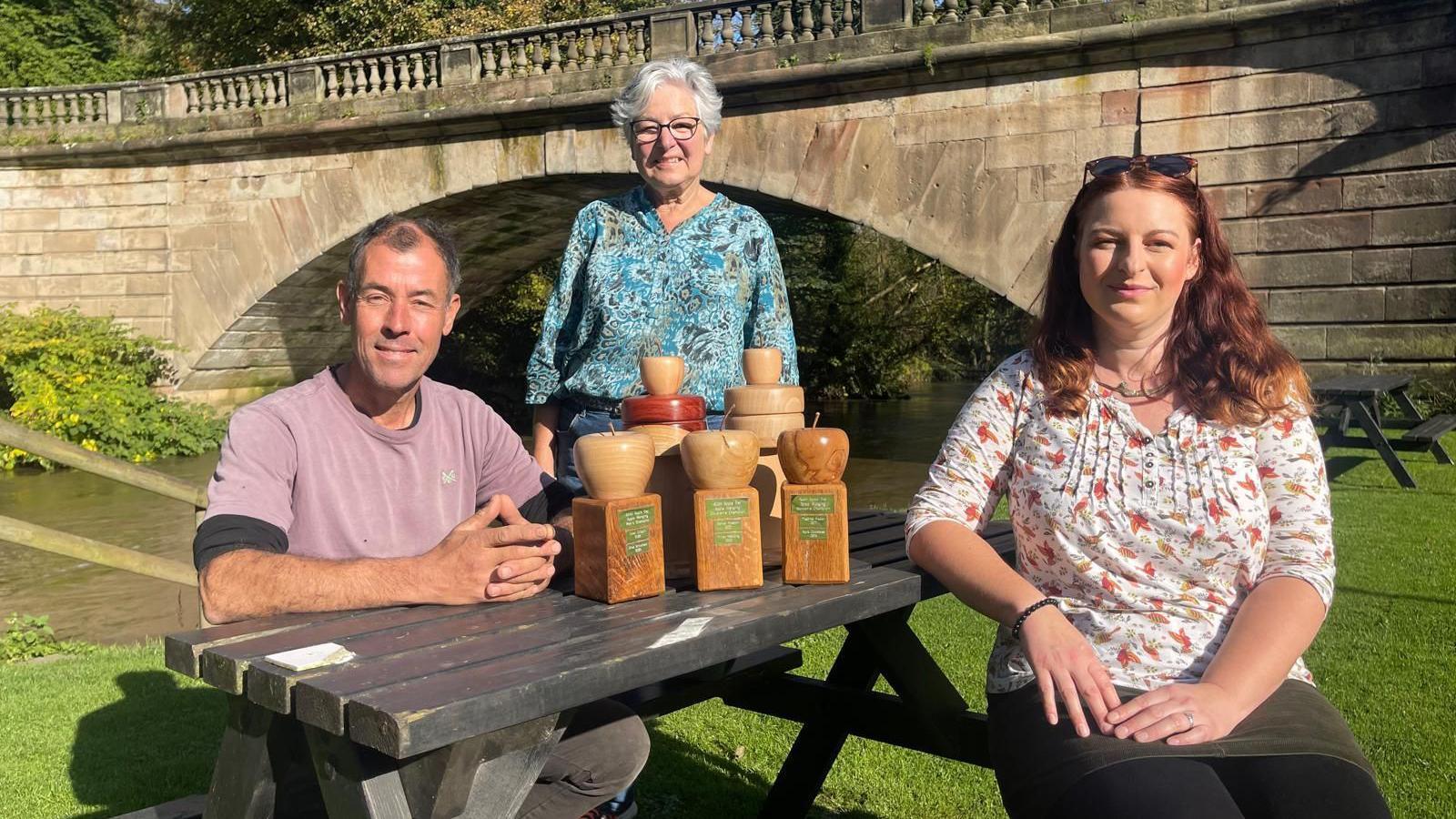 A wooden picnic bench sits on the grass beside a stone bridge. Sat at either side of the bench is a man in a lilac t-shirt and a woman in a flowery blouse. Stood at the end of the table is woman in a flowery green blouse, with short grey hair and spectacles. There are five wooden trophies between 25-35cm carved into an apple on a plinth.