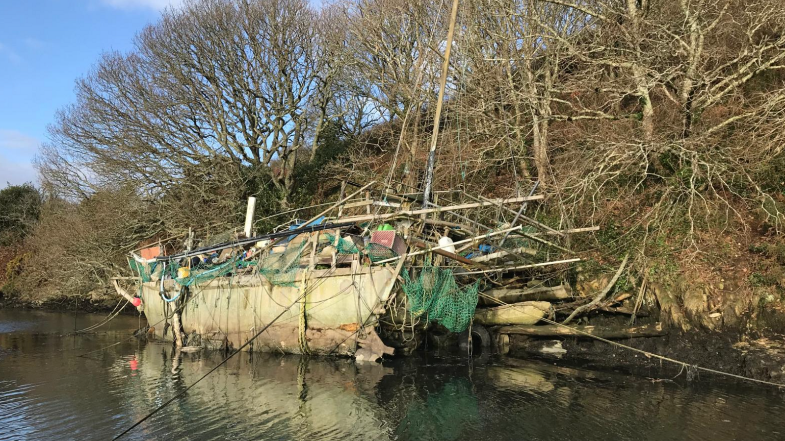 A dilapidated boat wreck on a riverbank with trees and bushes leading to the waterfront.