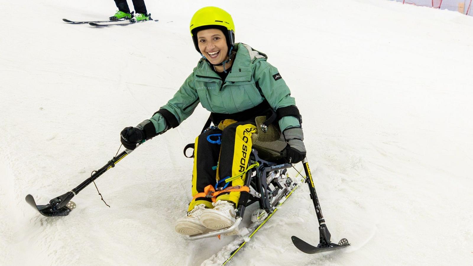 Ellie skiing wearing a yellow helmet, green ski jacket, yellow and black trousers and white trainers. She holds two ski poles out to either side and has a big smile