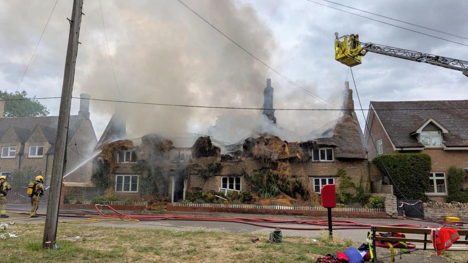 Firefighters at a large, two-storey thatched property. The roof has been destroyed. Only three chimney stacks remain standing. A plume of smoke is coming from the roof. A firefighter on the left is using a hose to douse the roof. An aerial ladder is at the top right of the picture. Several hoses are running along the road in front of the property.