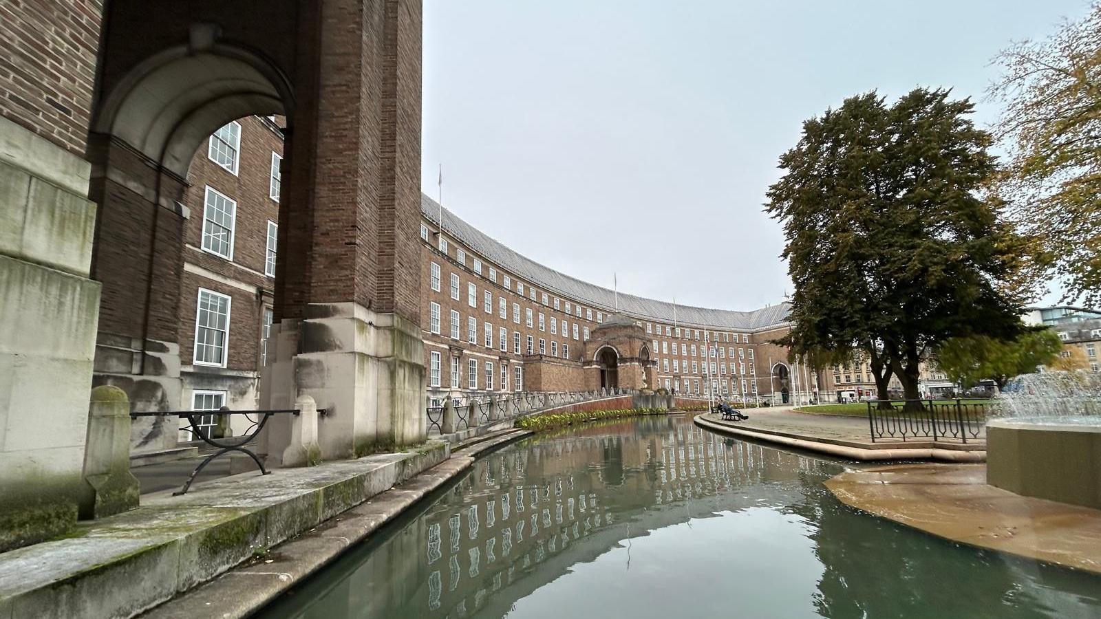 City Hall in Bristol. It is a grand, curved building with neatly lined windows and big pillars. There is water outside of it with a small fountain visible in the corner of the image.