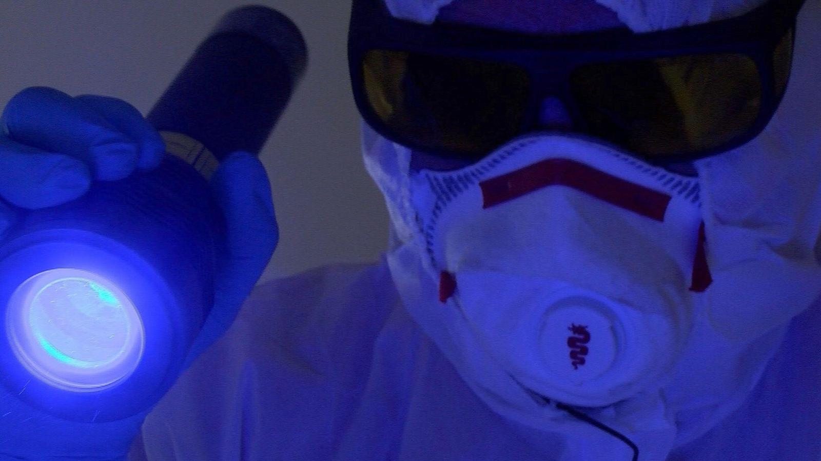 Jim Gildea is shown in full PPE, holding a torch emitting a blue light. He wears tinted lenses over his protective glasses as he looks for evidence of any remaining biological matter.