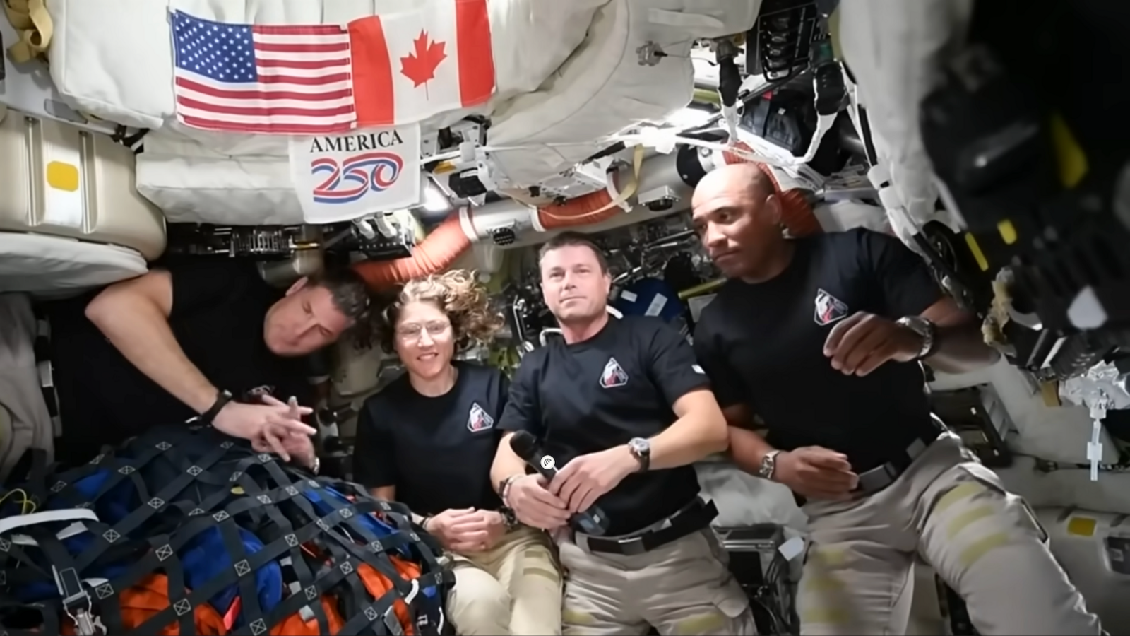 Inside a cramped spacecraft cabin, five astronauts float side by side in weightlessness. They wear matching black T‑shirts with a small mission patch on the chest and pale trousers with Velcro straps. Their faces are blurred, but their relaxed body language suggests they are chatting to camera. Behind them, every surface is crowded with white padded panels, cables, pipes and equipment. Above their heads, American and Canadian flags are pinned to a storage bag, with a colourful “America 250” sign between them. To the left, a netted bundle of blue and orange bags is tethered down, preventing it from drifting.

