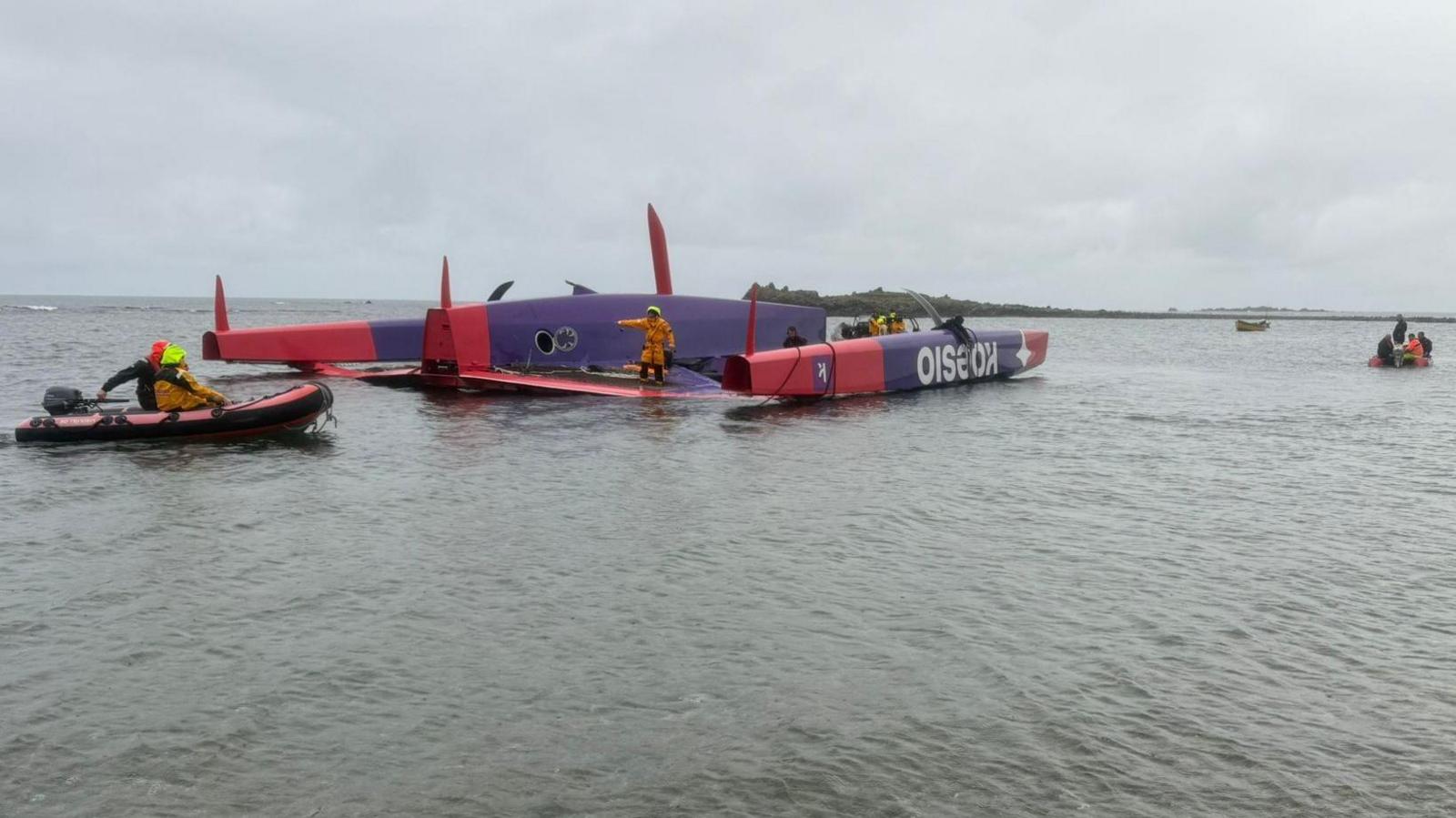 The image shows a large racing trimaran, overturned in shallow water. The hull is bright red and purple. Several support crew members in high-visibility gear are working around the vessel, using small inflatable boats to assist. The water is calm and shallow. The sky is overcast.