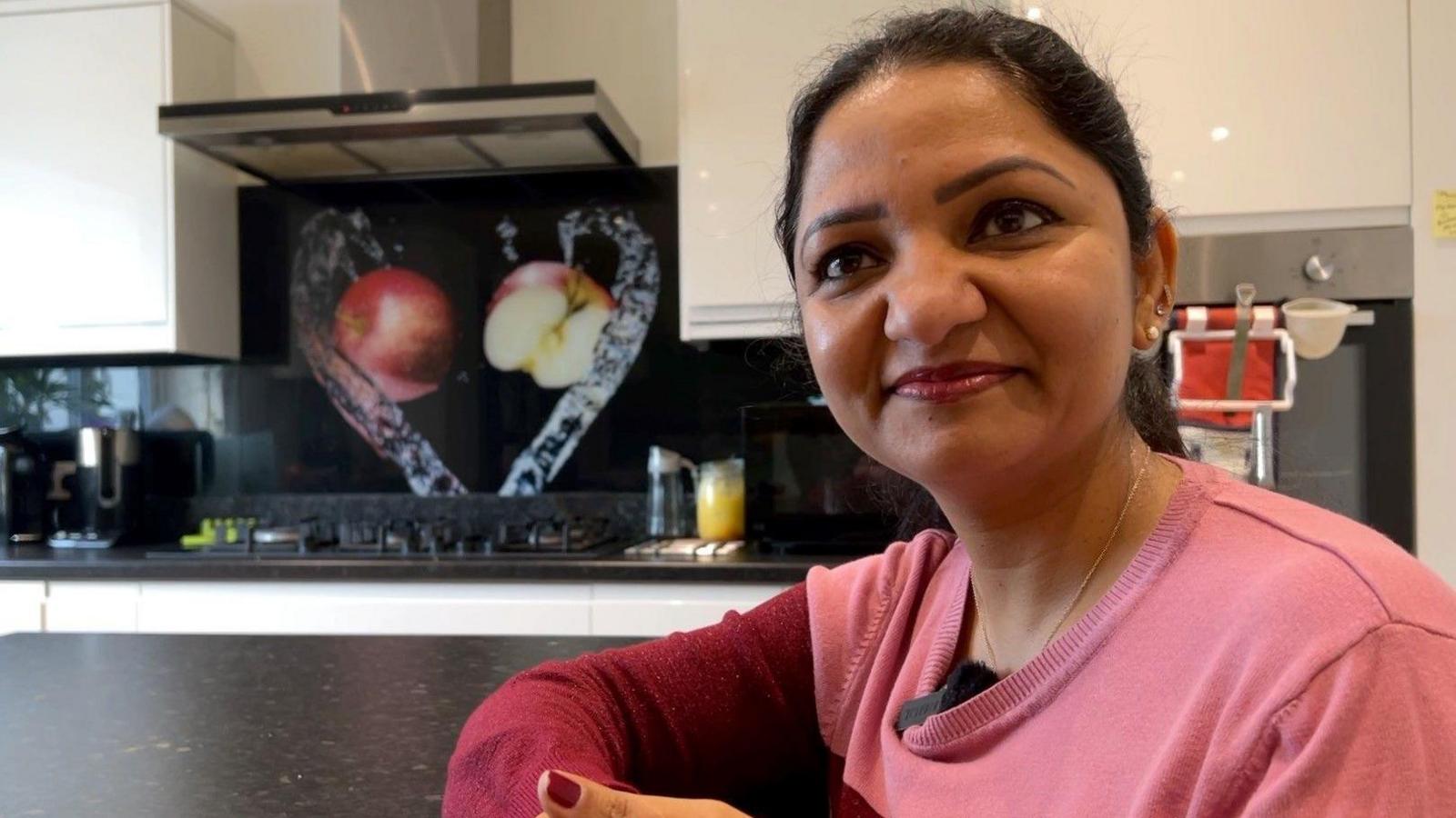 Sudha Sri Nukana sits at her kitchen table and faces the camera. She wears a pink jumper with darker pink sleeves and red lipstick.