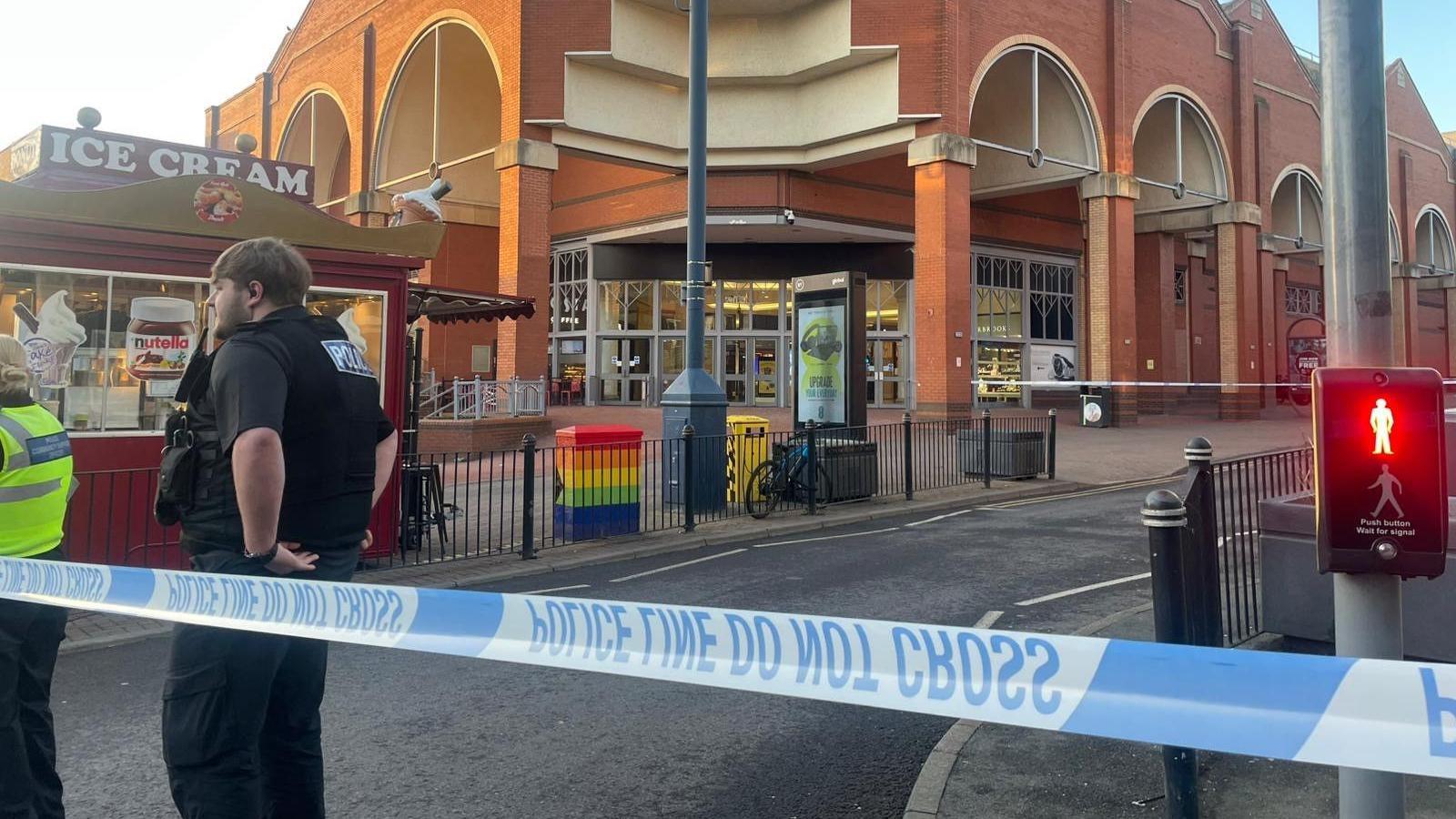 A police cordon around a shopping centre. There are two police officers seen standing by police tape and an ice cream stand is visible in the corner of the image.