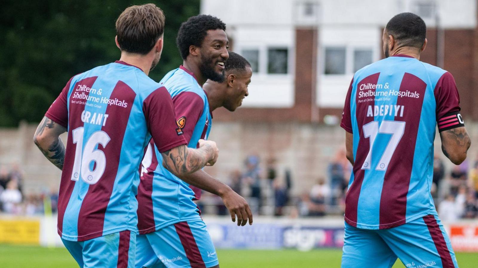 Devonte is smiling as he walks across a pitch with other Chesham United players. The players are wearing claret and blue striped kits. 