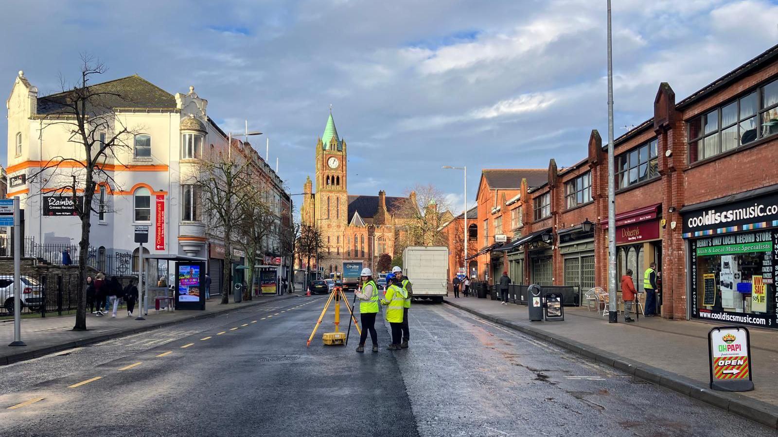 Three Ni Water workers stand in the middle of Foyle street in Derry. Either side of the street is lined with shops and cafes, and the Guildhall can be seen in the background. The workers are wearing high viz yellow jackets