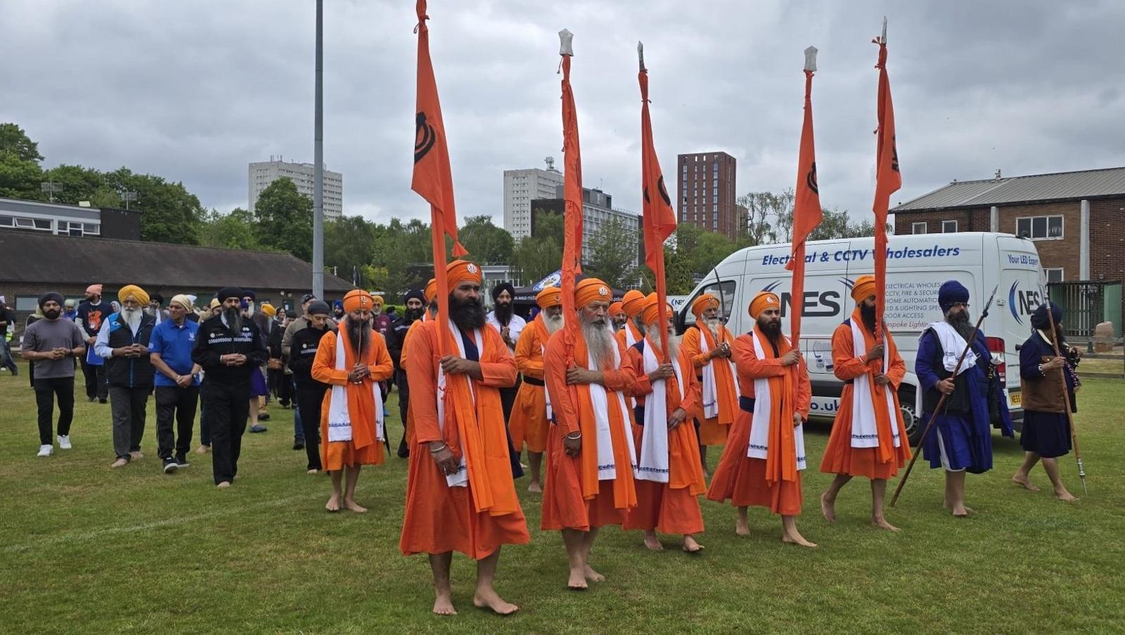 A group of people take part in a procession across a green lawn. The two front rows are made up of men wearing orange clothes and orange turbans. The five men at the very front are each holding an orange flag.