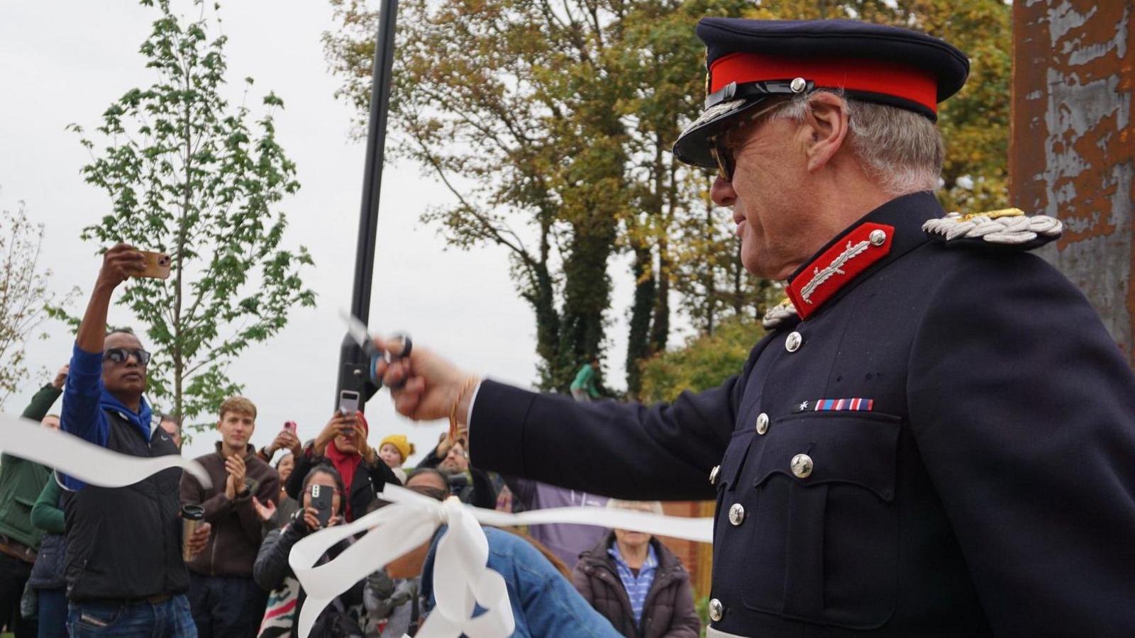 James Saunders Watson, the Lord Lieutenant of Northamptonshire, cutting a white ribbon, holding scissors in his hand, wearing a blue uniform, with a hat. He looking to the left. A crowd of people are in front of him, holding phones and clapping. Trees can also be seen.