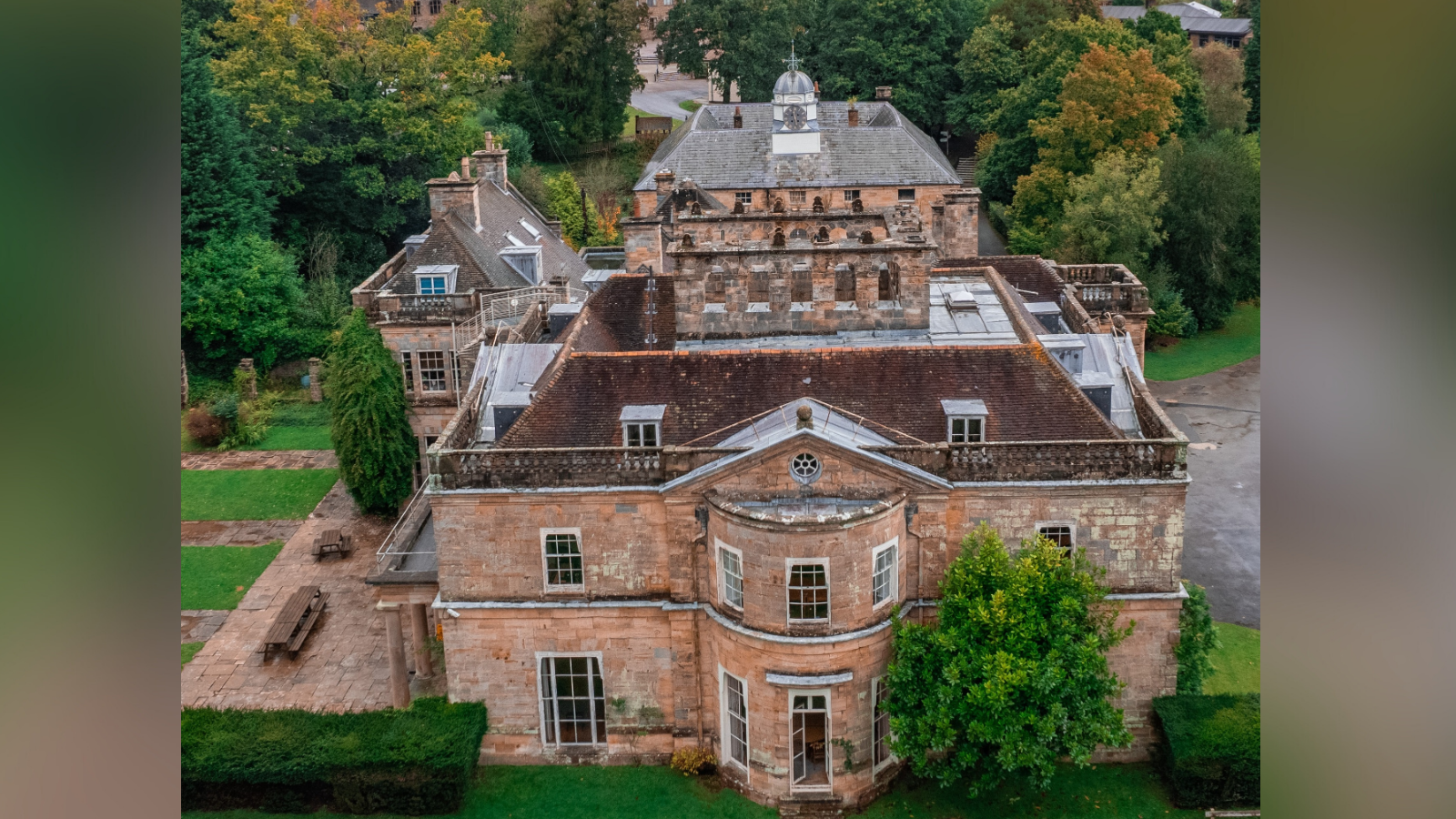 An aerial picture of Michael Hall Waldorf School. The front building which extends further back is visible, as is a side building to the left. Benches can be seen to the left-hand side of the building, which is encased by a green hedge. There are a number of trees behind the buildings.