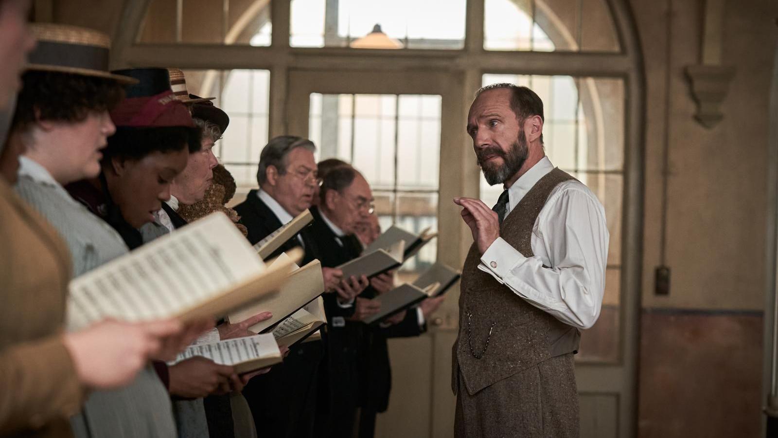 A group of people in historical costume stand in a line, holding sheet music, mid-rehearsal. A man, Ralph Fiennes, in a white shirt and waistcoat gestures with one hand, likely conducting.