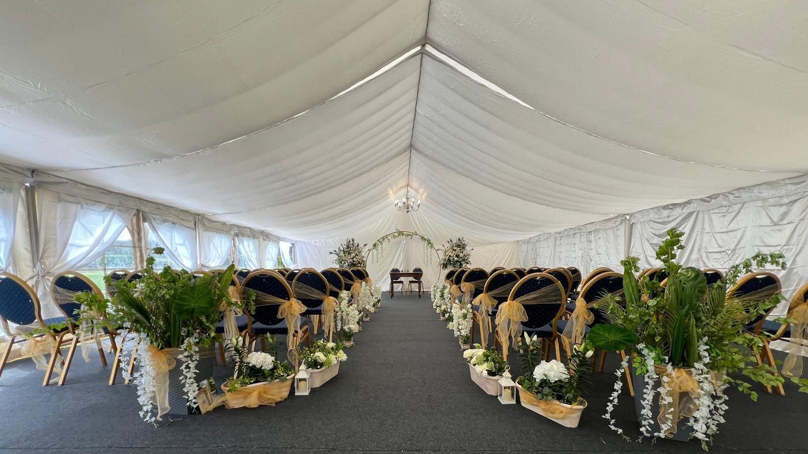 The inside of a large marquee with decorated chairs alongside a central aisle, ready for a wedding to be held