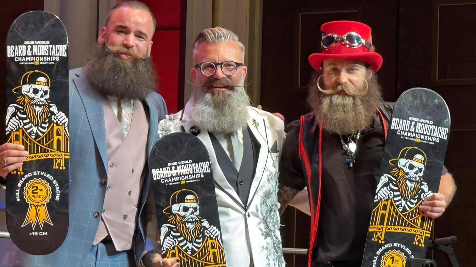 Three men with elaborate beards and moustaches stood arm-in-arm on a stage. The man on the left is wearing a light blue checked suit with a mauve-coloured waistcoat and a floral-patterned tie. He is holding a skateboard featuring a black and yellow design and a second place ribbon badge. The man in the middle has short grey hair and is wearing black glasses and an off-white suit with a grey waistcoat and a light blue tie. He is also holding a skateboard. The man on the right - Lewis Aylmer - is wearing a red top hat and a black and red outfit. He is holding a skateboard with a fist place ribbon on.