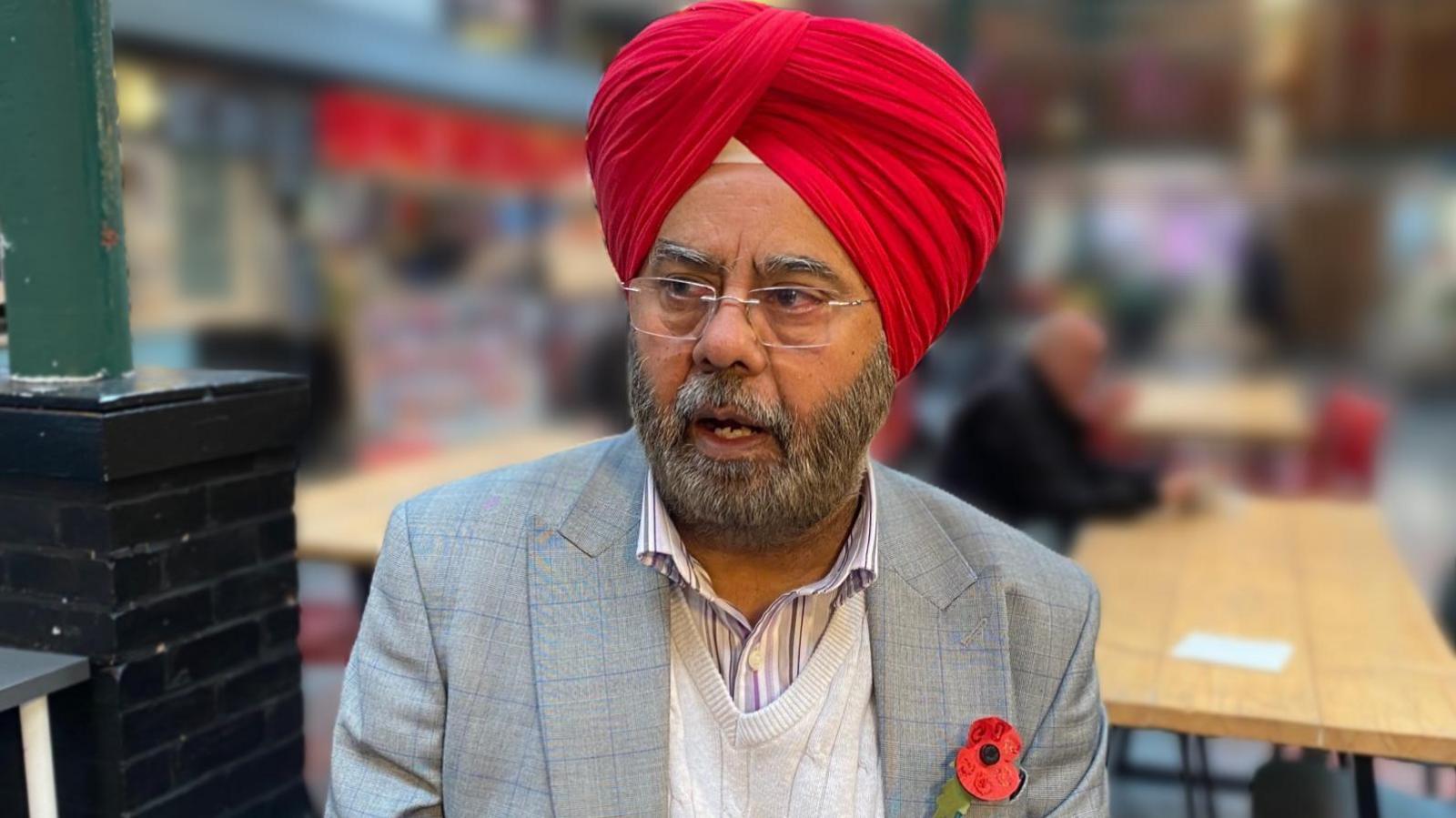 A bearded man wearing a grey blazer, cream jumper and red turban. He is also wearing a poppy badge. In the background is a green steel column and wooden benches.