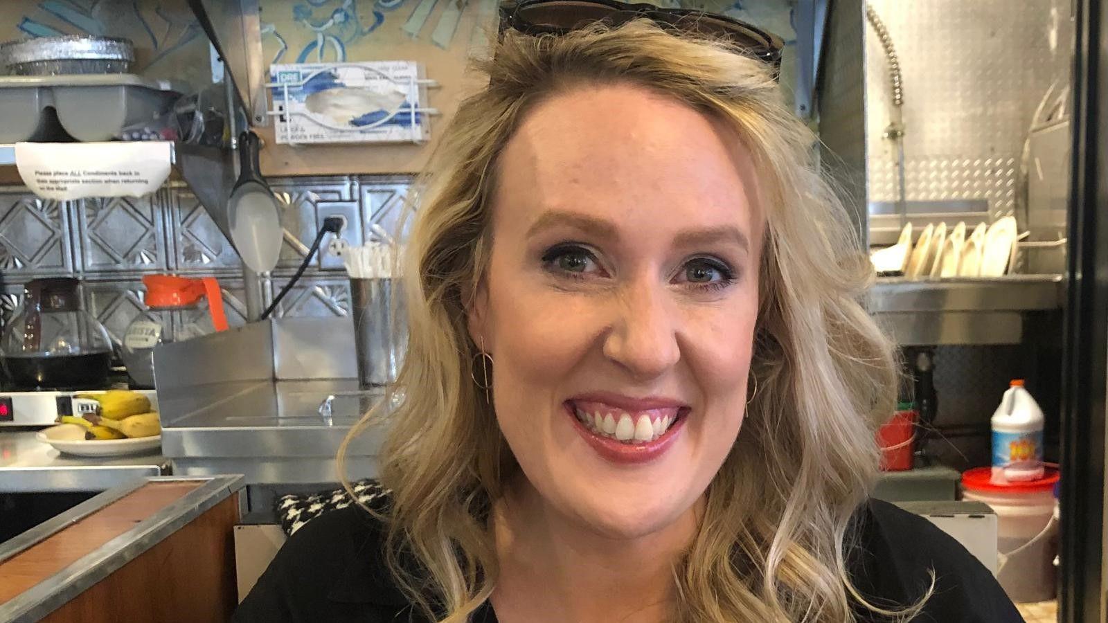 Catherine Thom with blonde wavy shoulder length hair and blue eyes smiling at the camera wearing a black top in a cafe with the kitchen seen behind her