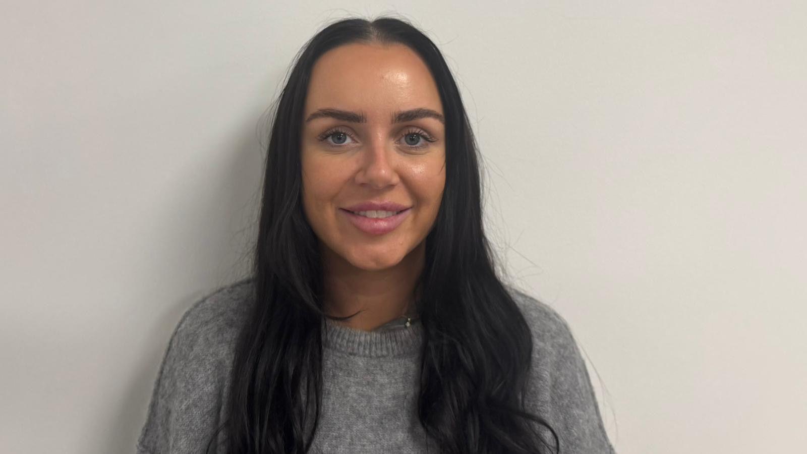 A girl with dark brown hair stands against a white wall. She is smiling, teeth showing as she wears a grey woollen jumper.