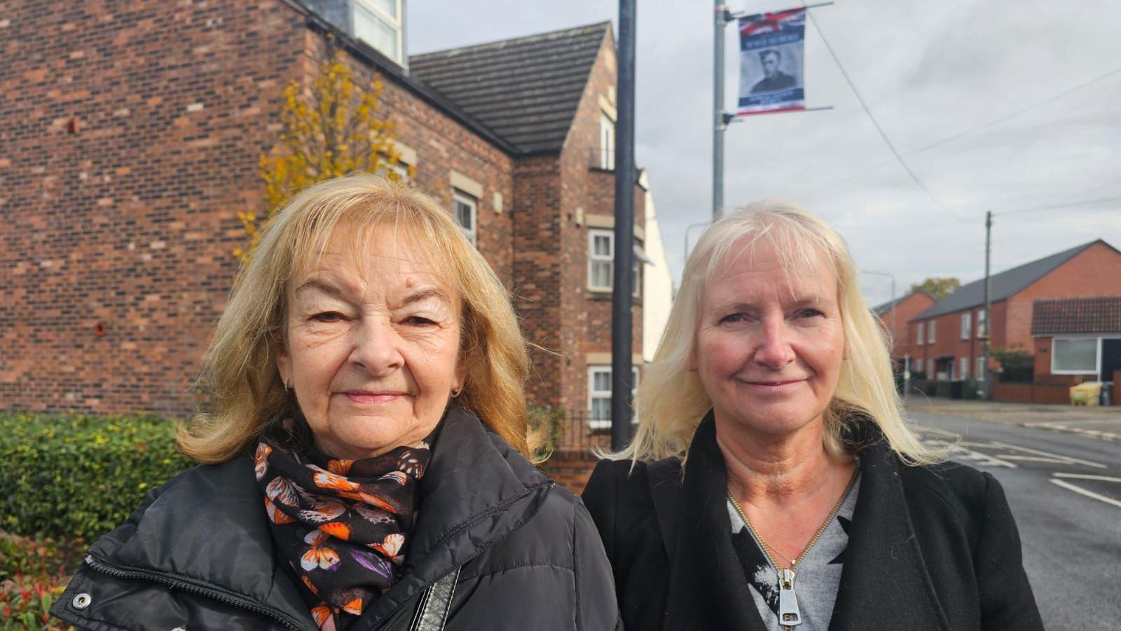 Jean Thompson (left) has blonde short hair with a fringe and is wearing a black coat with a colourful butterfly printed scarf. Her sister Lynda (right) is wearing a black coat and a grey jumper. She has blonde hair. The pair are standing at a roadside and in the background you can see a lamppost with a picture of their uncle on it.