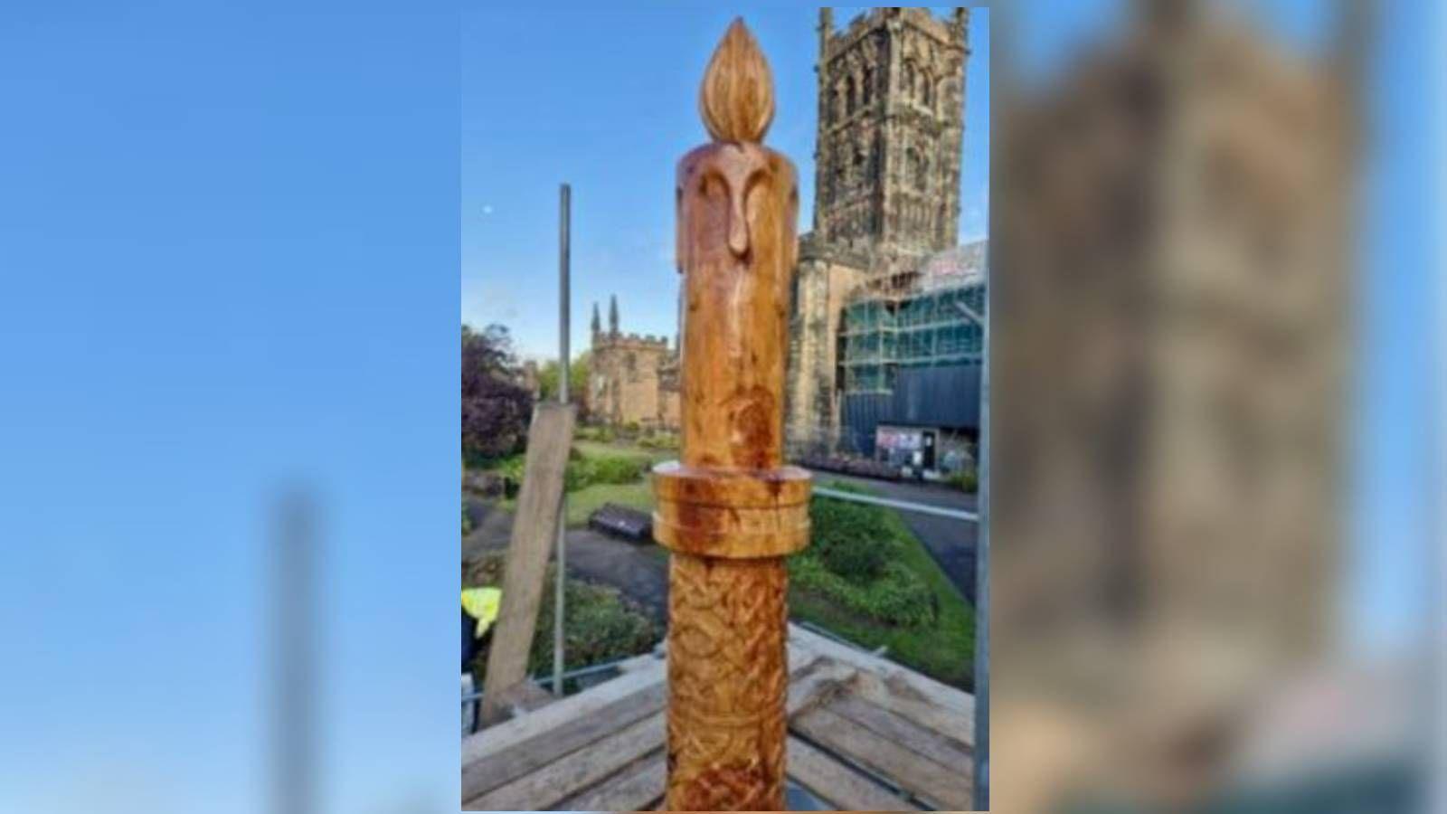 A wooden carved sculpture depicting a candlestick, candle and flame, made from cedar wood. It stands in the grounds of a church, which can be seen in the background.