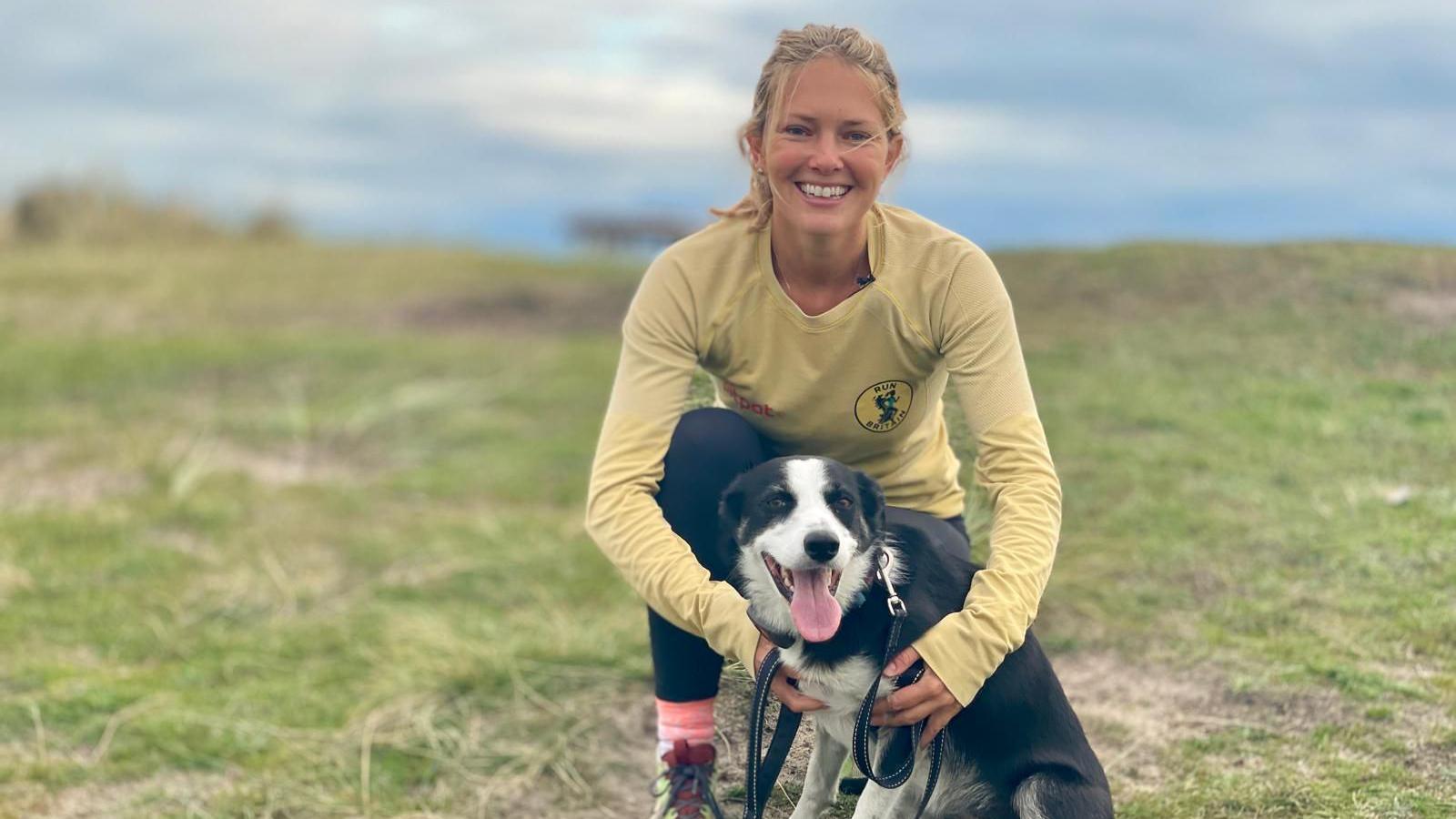 Megan Boxall was bending down on one knee and looking towards the camera smiling. She had blonde hair in a ponytail and was wearing a yellow long sleeved top with a logo. She was holding a black and white medium sized dog in front of her.