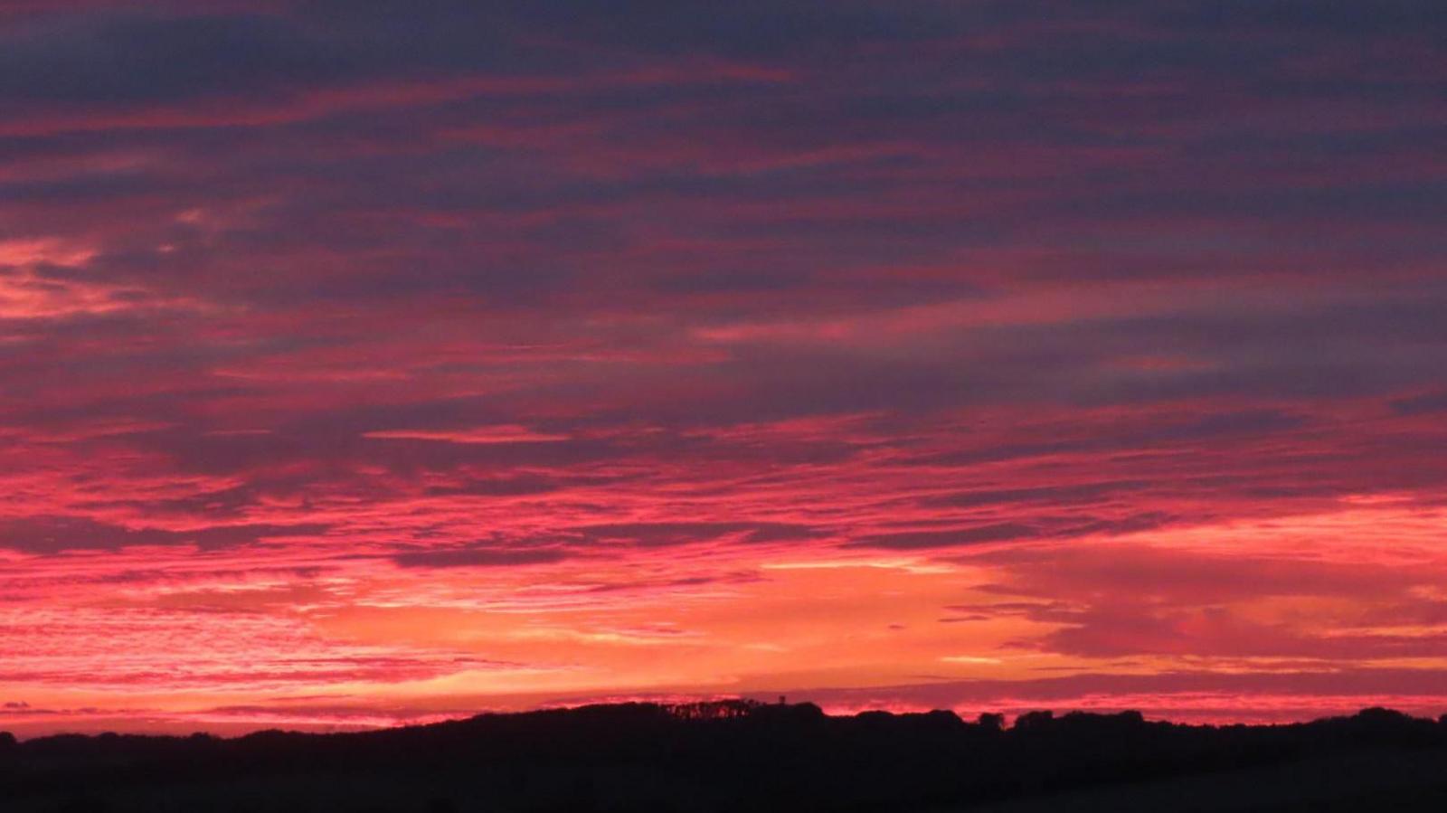 A sky of vibrant red, pink and orange hues mixed with bits of thick, blue cloud stretching horizontally across the top of the skyline. At the bottom, the silhouette of hills is visible.