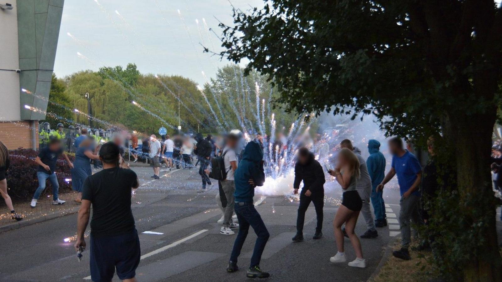 A large group of people in the middle of a road, there is an explosion that looks like a firework going off. Towards the side police in helmets and hi-viz jackets are visible