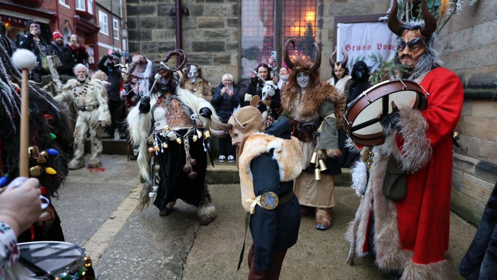 A group of people dressed up for Whitby Krampus Run festival perform in the street, wearing horns, fur and masks and banging drums 