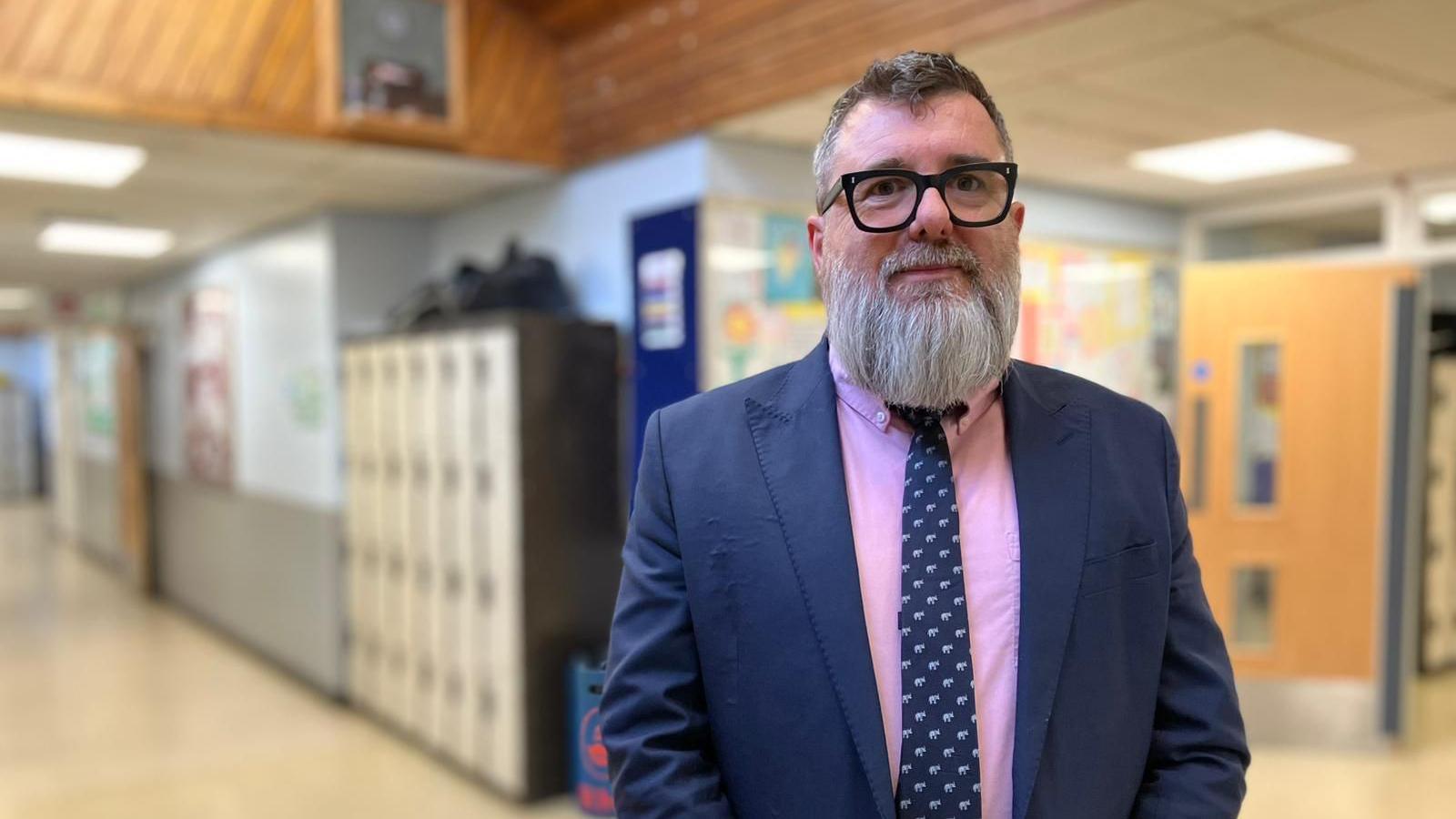 Head teacher Richard Horsfield standing in the corridor of his school. He is wearing a blue suit and tie with a pink shirt, and has black glasses and a beard.