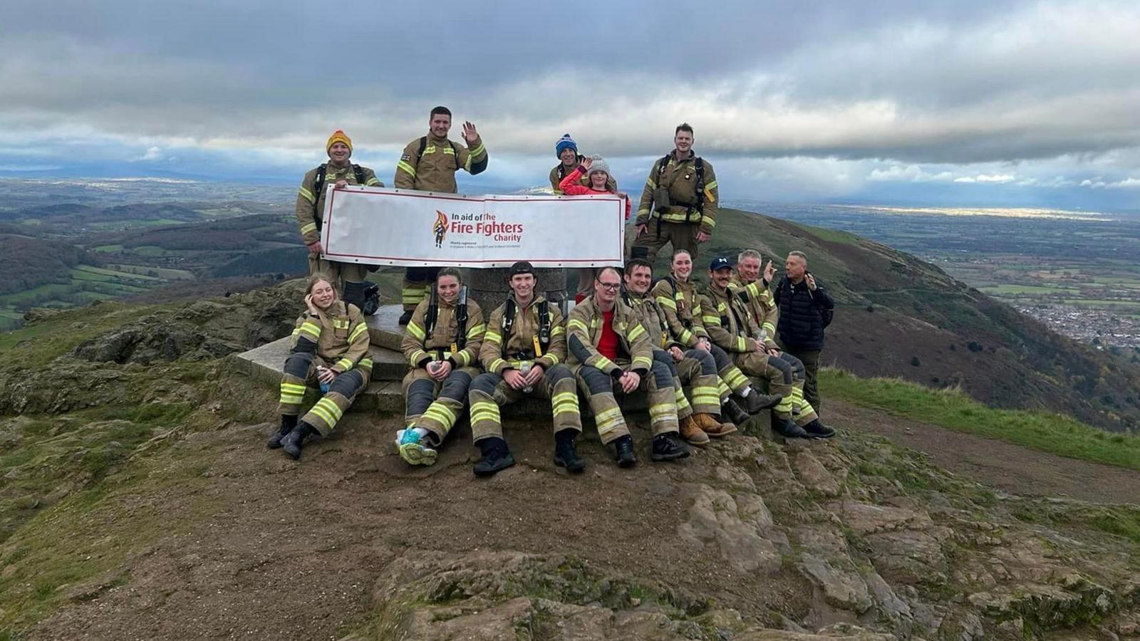 A group of people in firefighting uniform photographed at a summit trig point, carrying a Fire Fighters Charity banner.