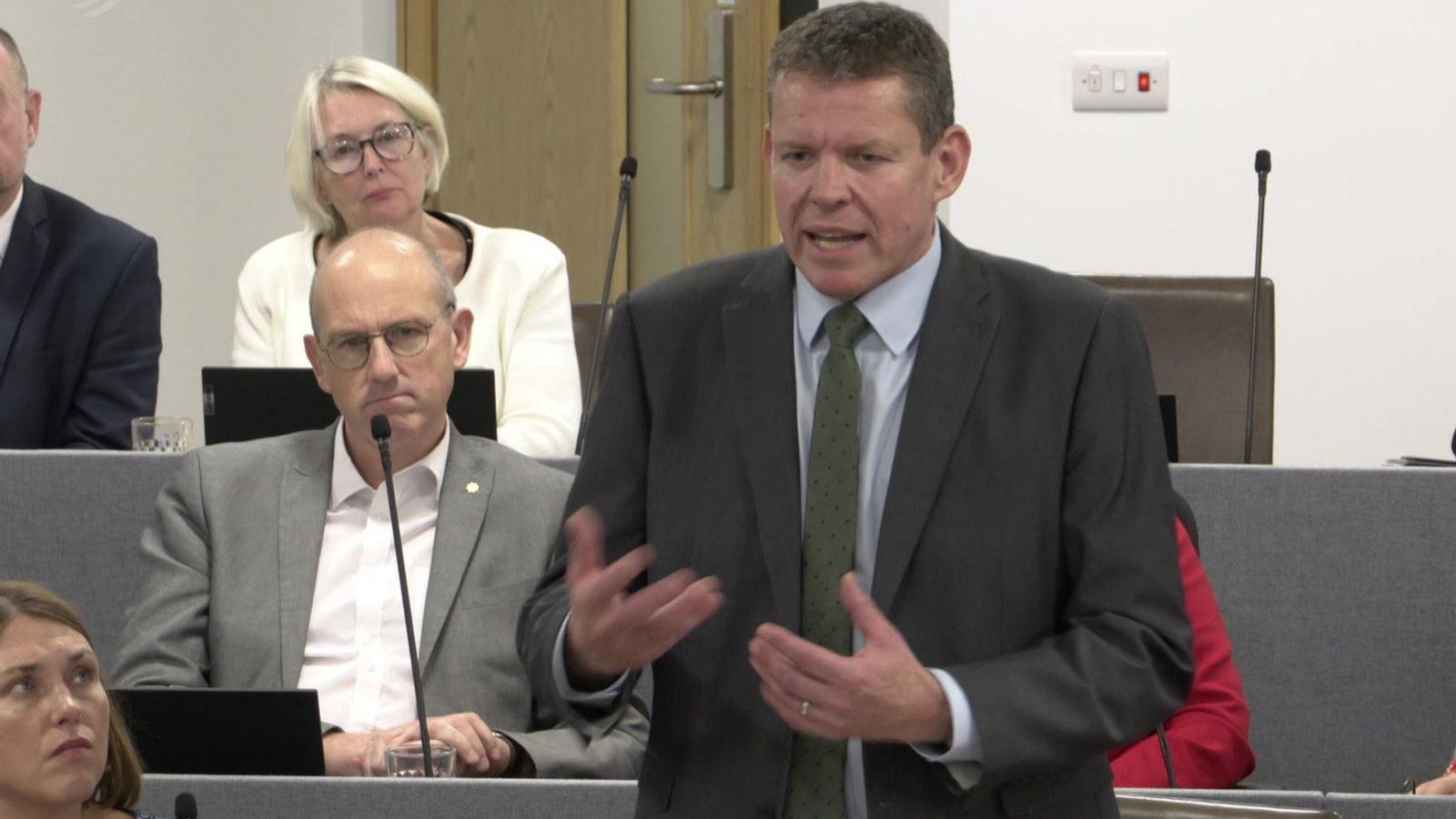 Rhun ap Iorwerth addressing Eluned Morgan during First Minister's Questions. He is gesturing with his hands and has Plaid Cymru colleagues behind and to the side of him.
