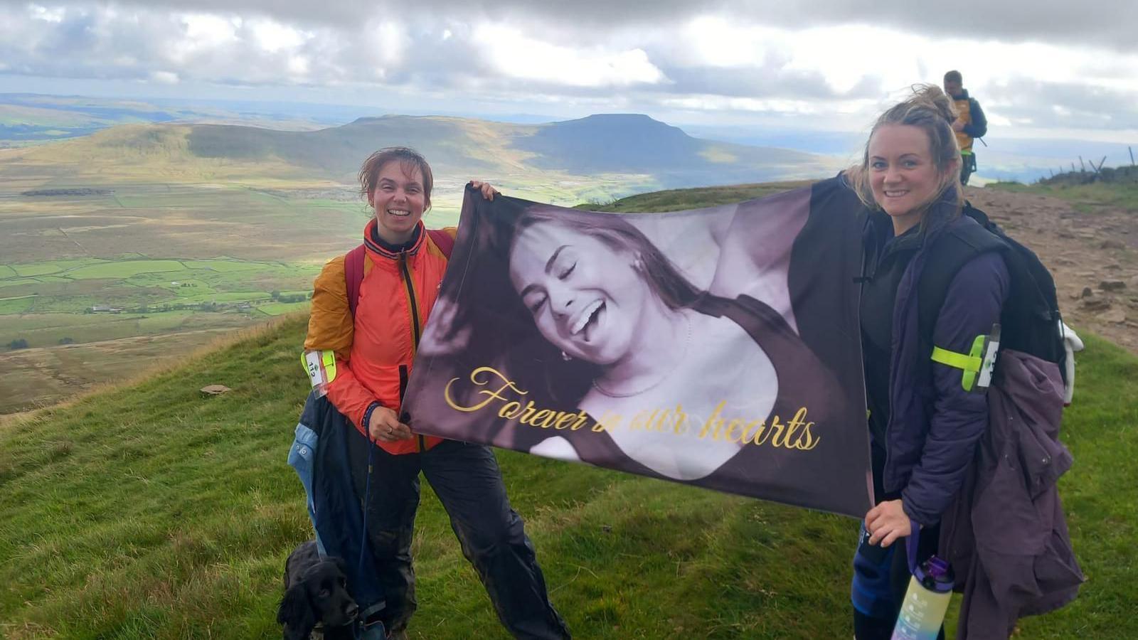 Two women are holding a big picture of Livia Wilson as they stand at the peak of one of the Yorkshire Three Peaks. The view behind them is pretty, with clear blue skies and hills.   
