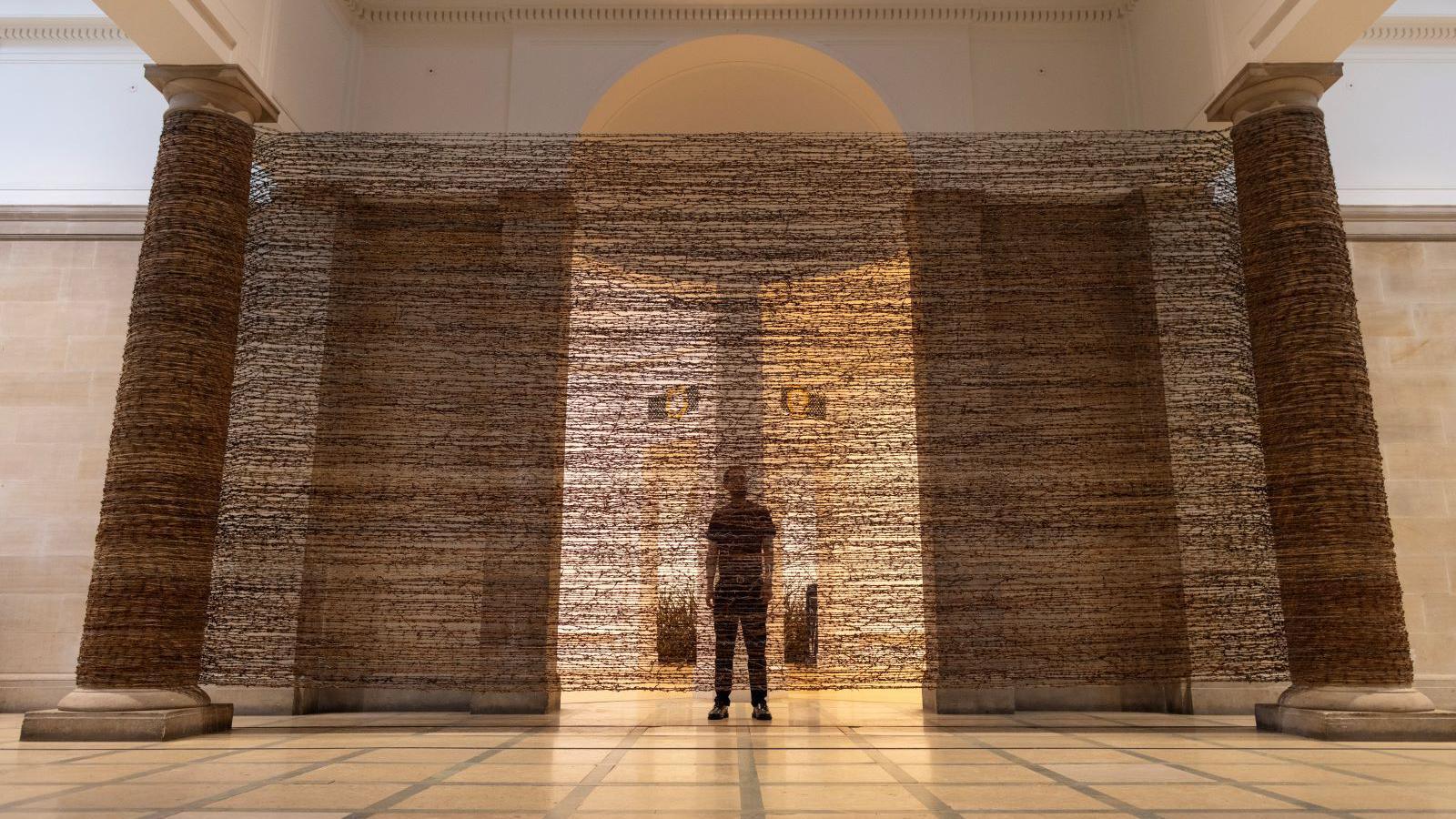 A mesh fabric made of rusted barbed wire stretches between two pillars with a man silhouetted behind it.