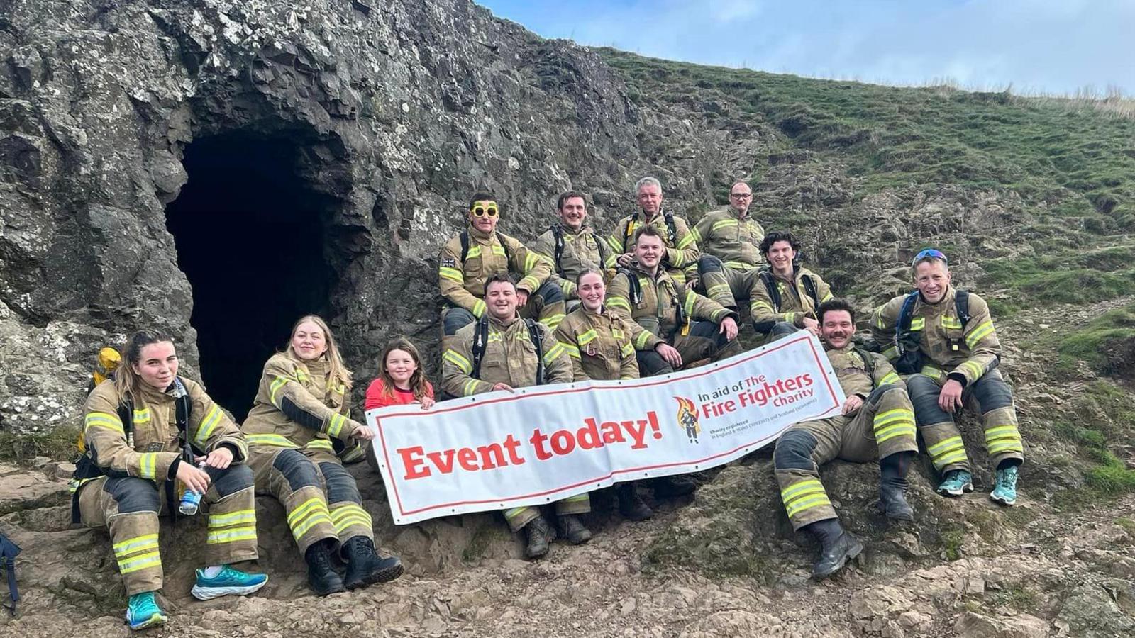 A group of 12 men and women in khaki and hi-viz firefighting uniforms, along with a young girl. They are sitting on rocks on a hillside, and holding a banner reading "Event today in aid of the Fire Fighters Charity".