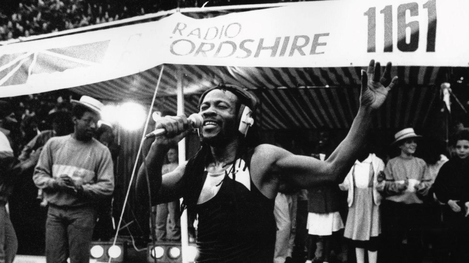 The Man Ezeke singing into a microphone in front of a tent outside. Behind him people are joining in with the music. He has his arm in the air and his eyes closed.