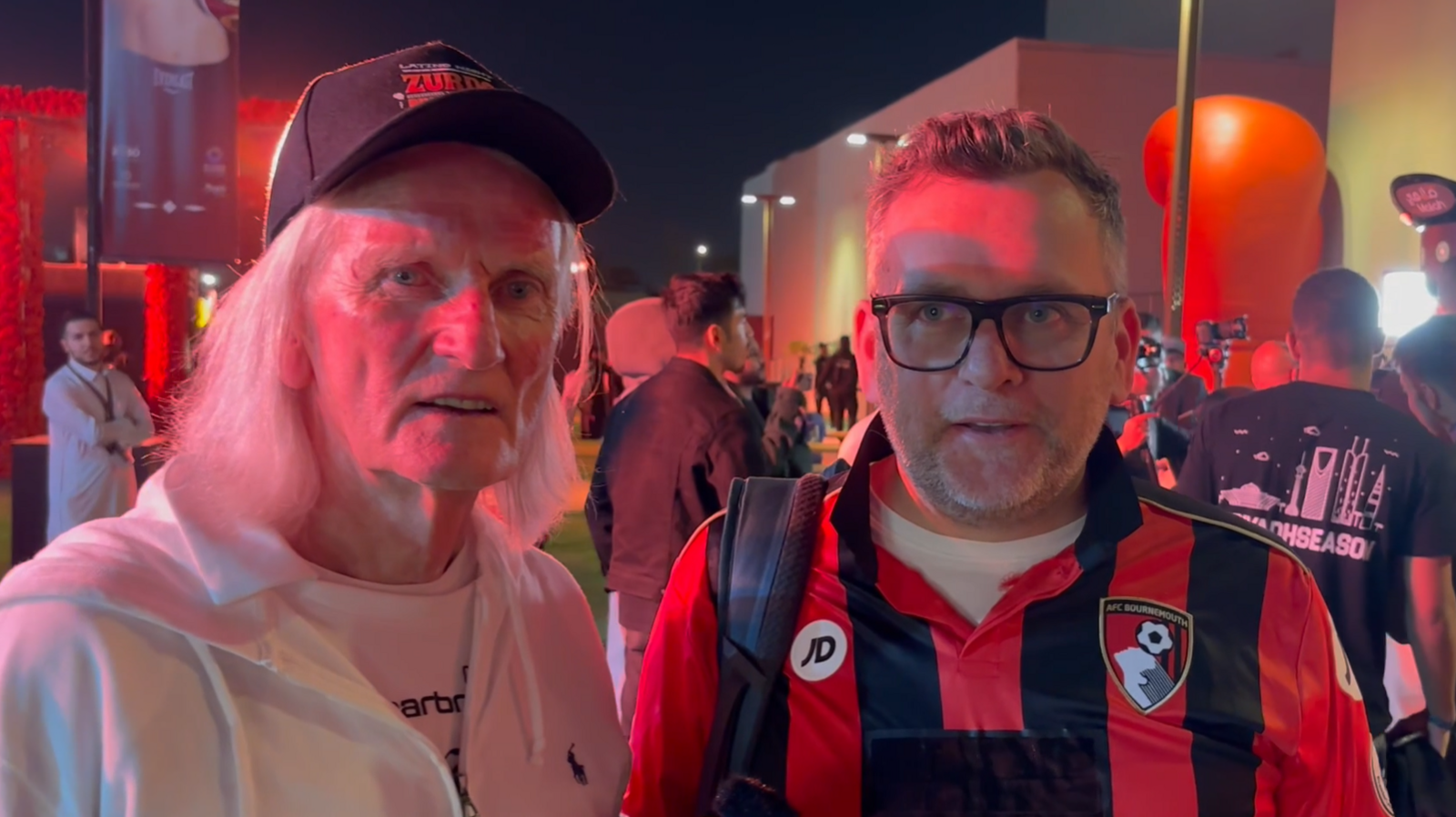 A man in a white hoodie and black baseball cap and a man wearing a red and black stripy shirt looking at the camera outside a busy stadium.