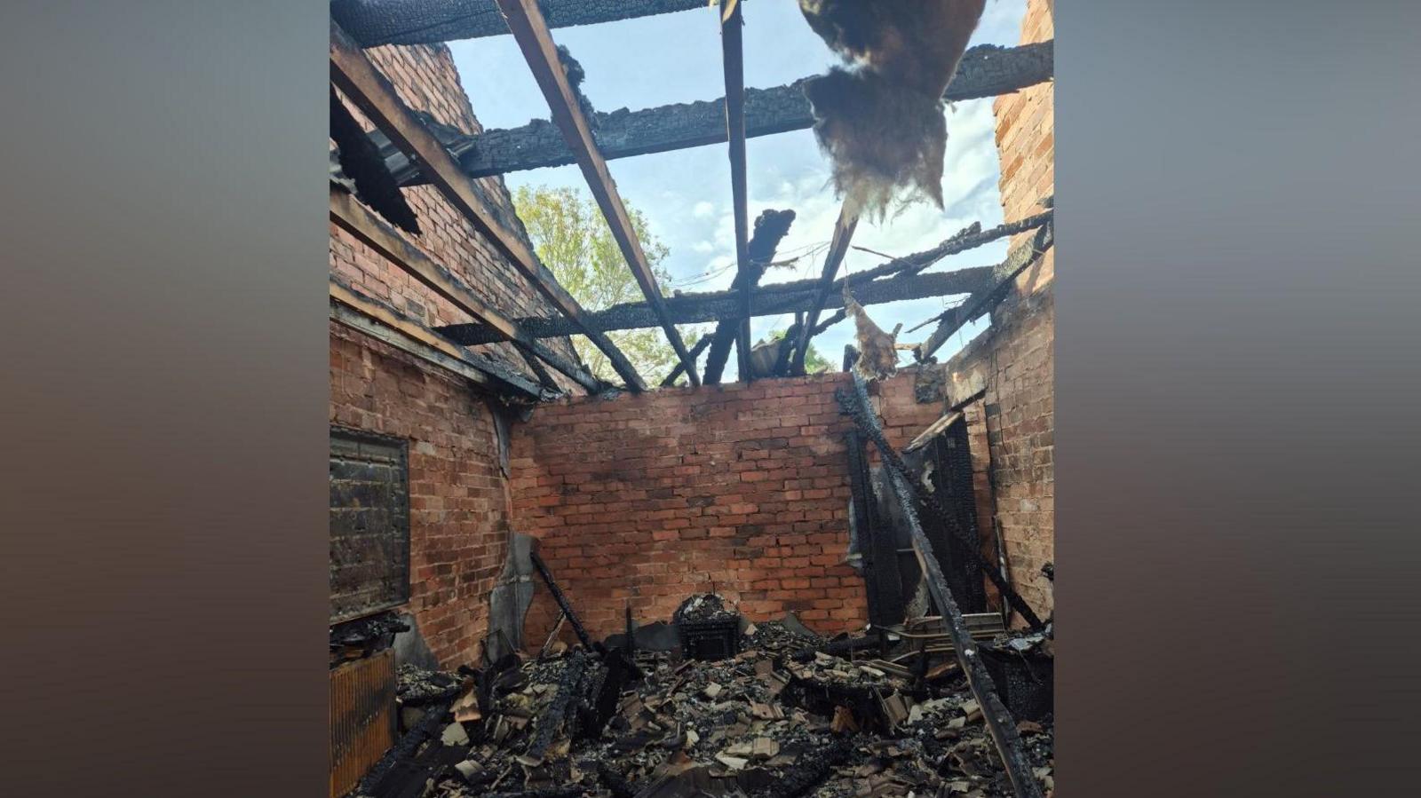 A damaged house including a missing roof and lots of damaged, burnt items on the floor and an exposed brick wall.