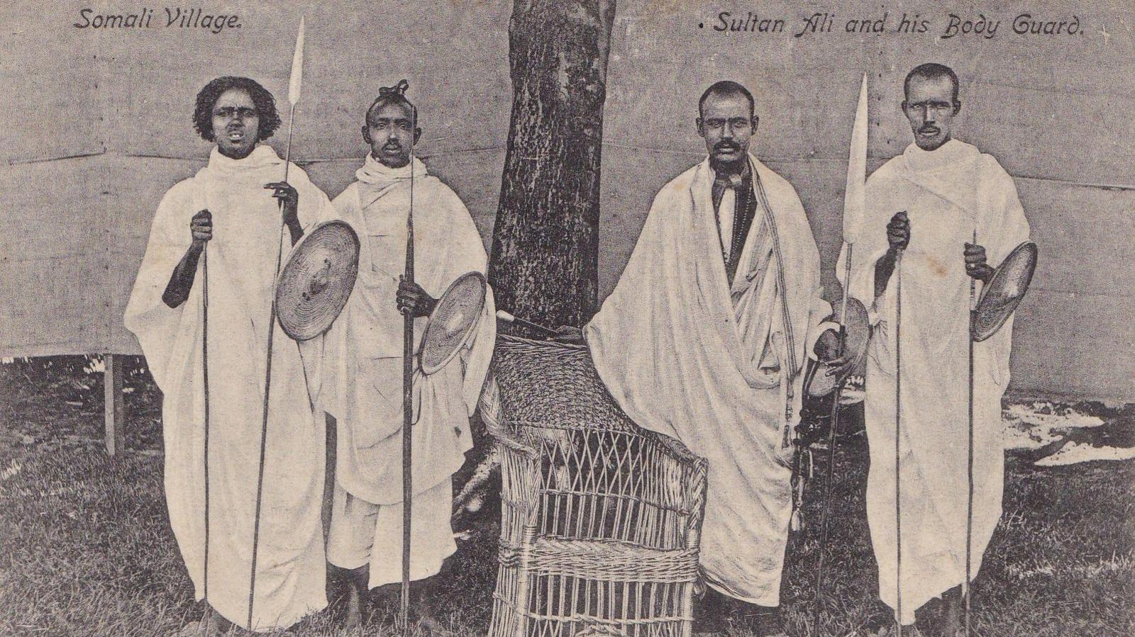 A group of four black men wearing white robes in a traditional African style stand in front of a tree. They hold spears and shields. The faded black and white photo is marked 'Somali Village - Sultan Ali and his bodyguard'.