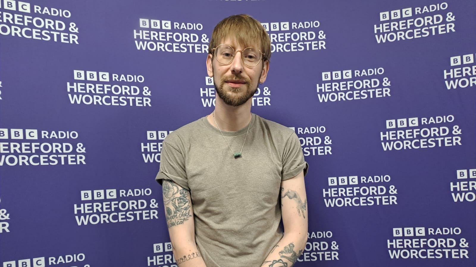 Man wearing round glasses, a grey t-shirt and green necklace is standing in front of a purple wall with the words "BBC Radio Hereford and Worcester" in the background.