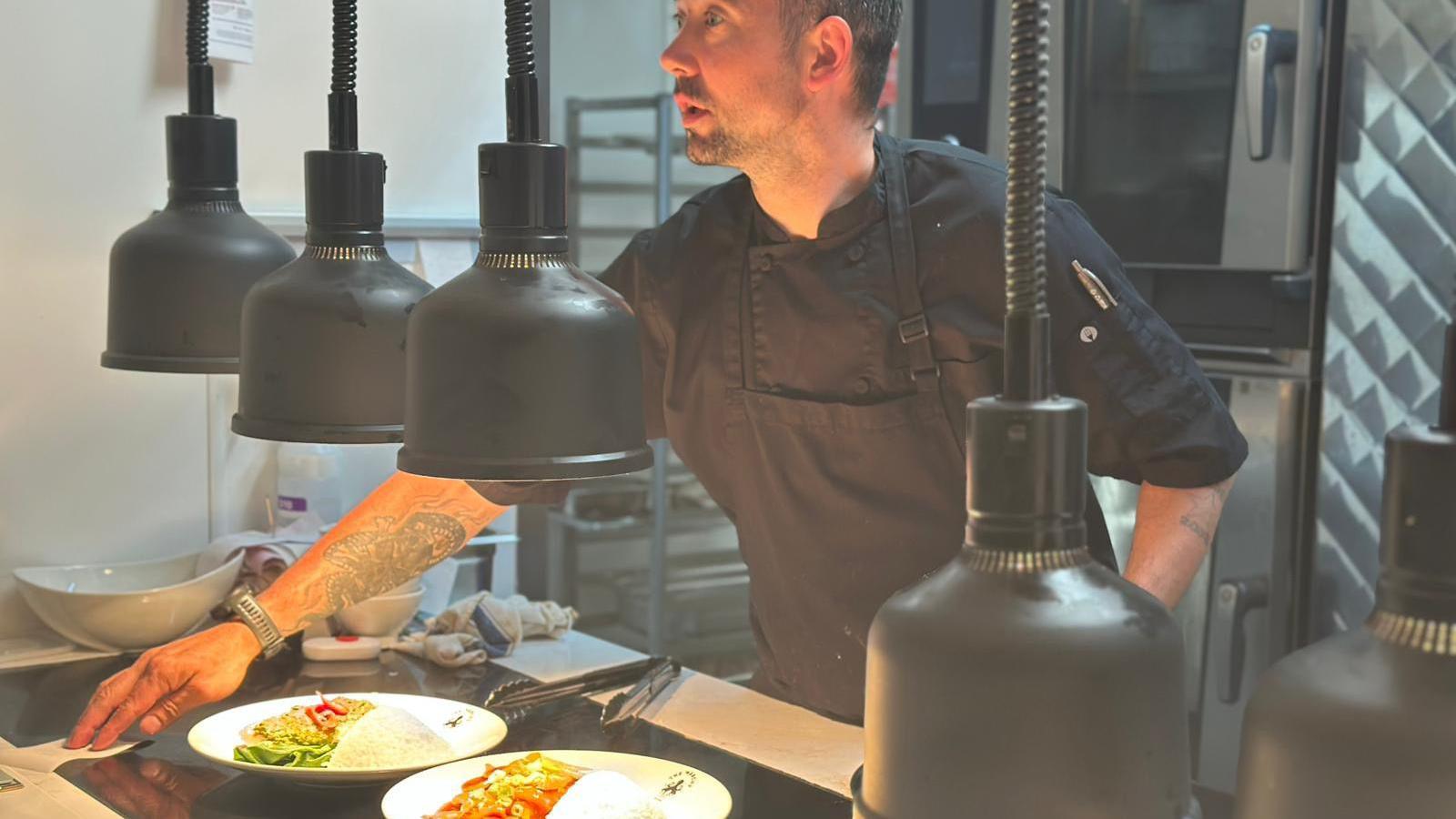 A man is serving food in a kitchen. There are two plates of food on a black counter. The man is wearing a black shirt and apron. There are large black lights hanging down. The background is of kitchen apparatus.